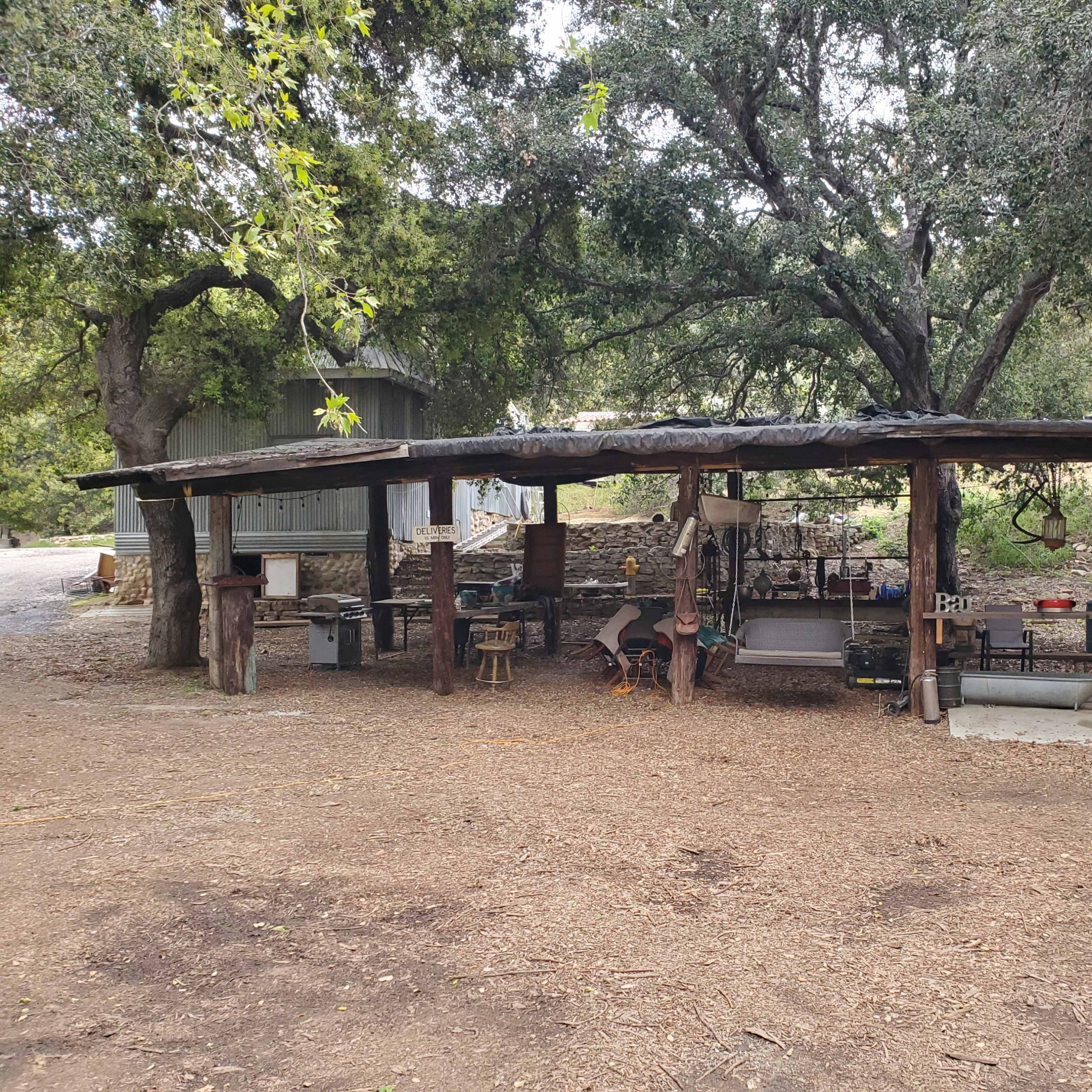 An outdoor cooking area with a roof supported by wooden posts, surrounded by trees and dirt ground.