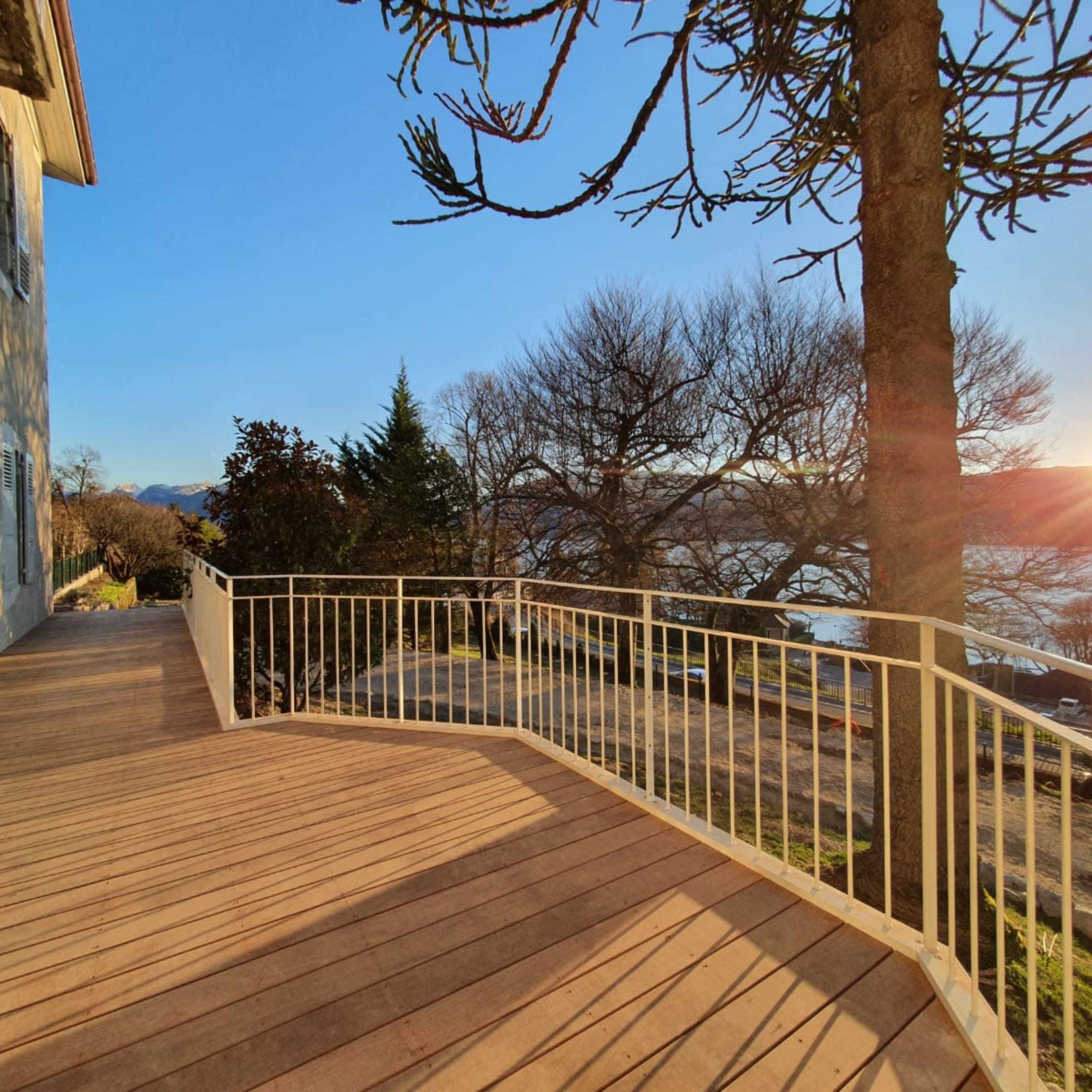 A wooden deck beside a lake, with a house on the left and a sunset visible in the background.