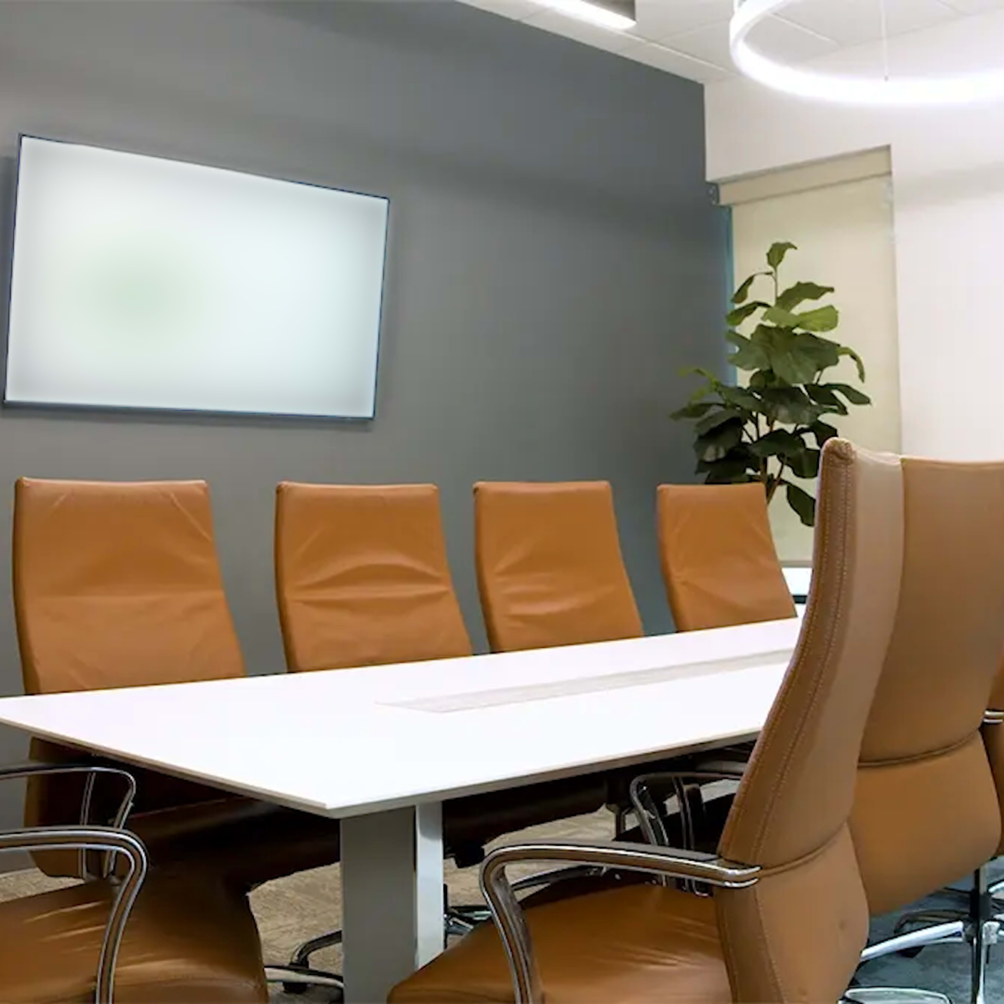 A modern conference room features a long white table surrounded by leather chairs and a large screen mounted on the wall.