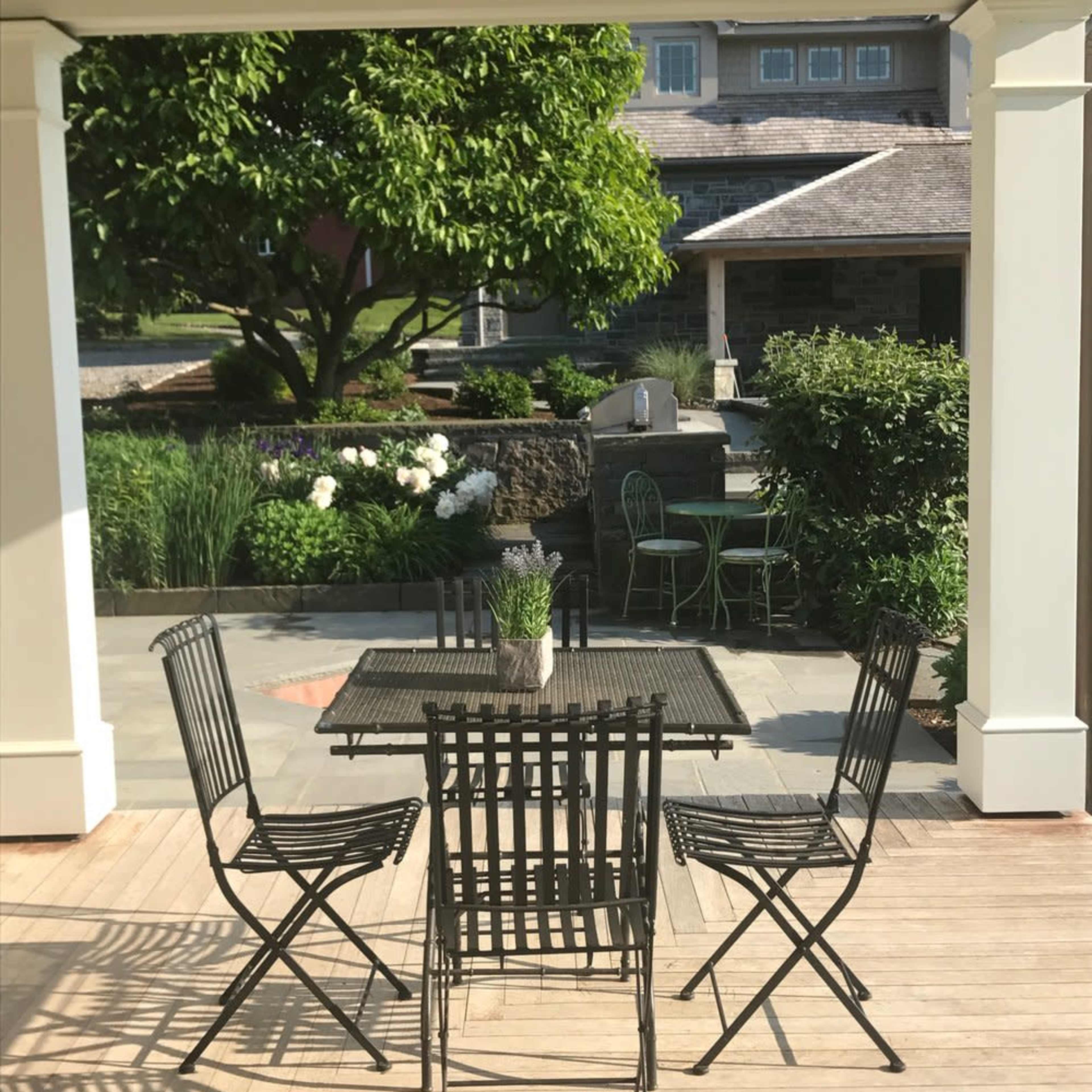 A patio area features a table surrounded by four black metal chairs, with greenery and a garden visible in the background.