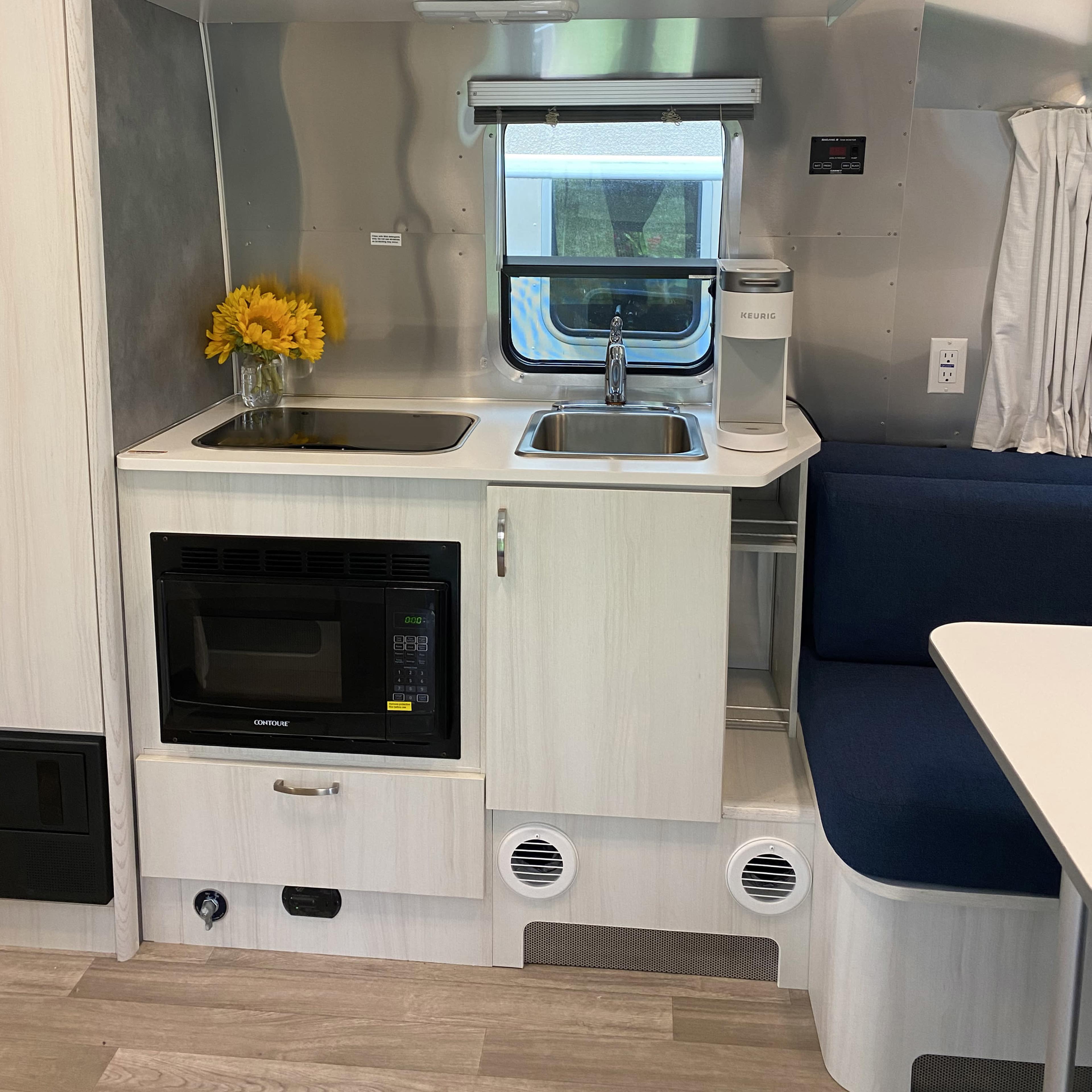 A compact kitchen area inside a trailer, featuring a microwave, a sink with a faucet, and a blue seating area.