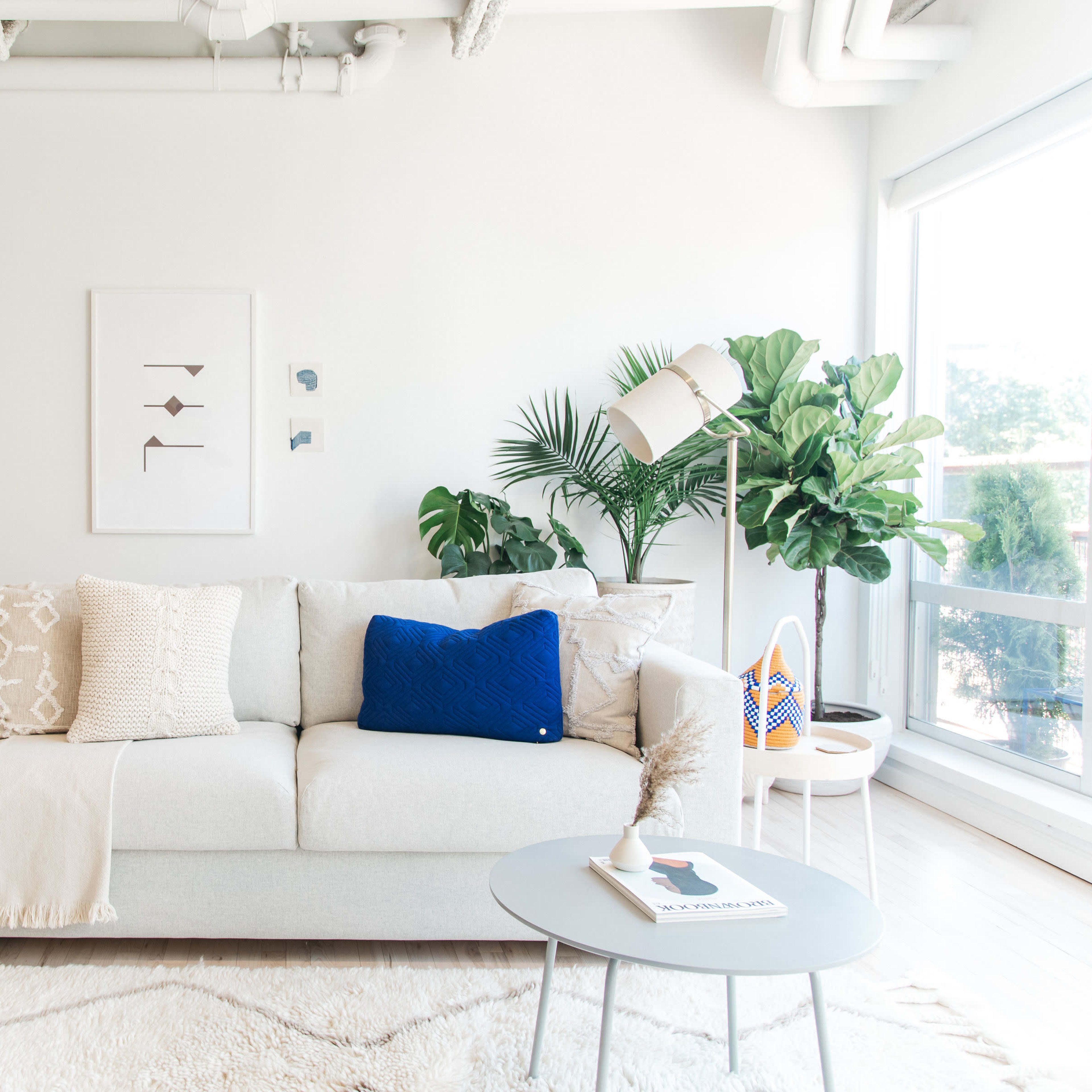 A bright living room features a light-colored sofa with decorative pillows, a small coffee table, and potted plants, complemented by minimalist decor on the walls.
