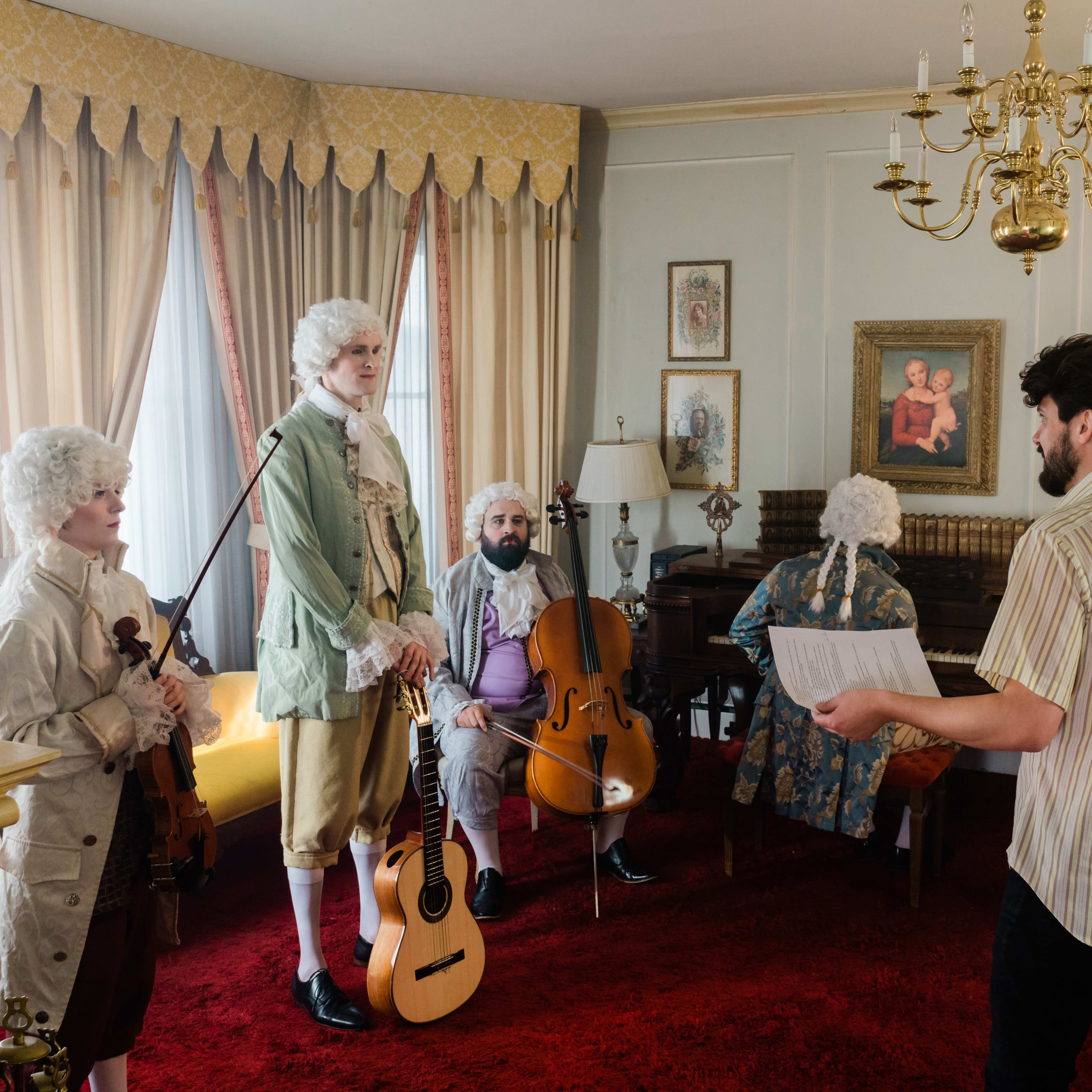 A group of musicians dressed in period costumes plays instruments in a styled room while a man stands holding a script.