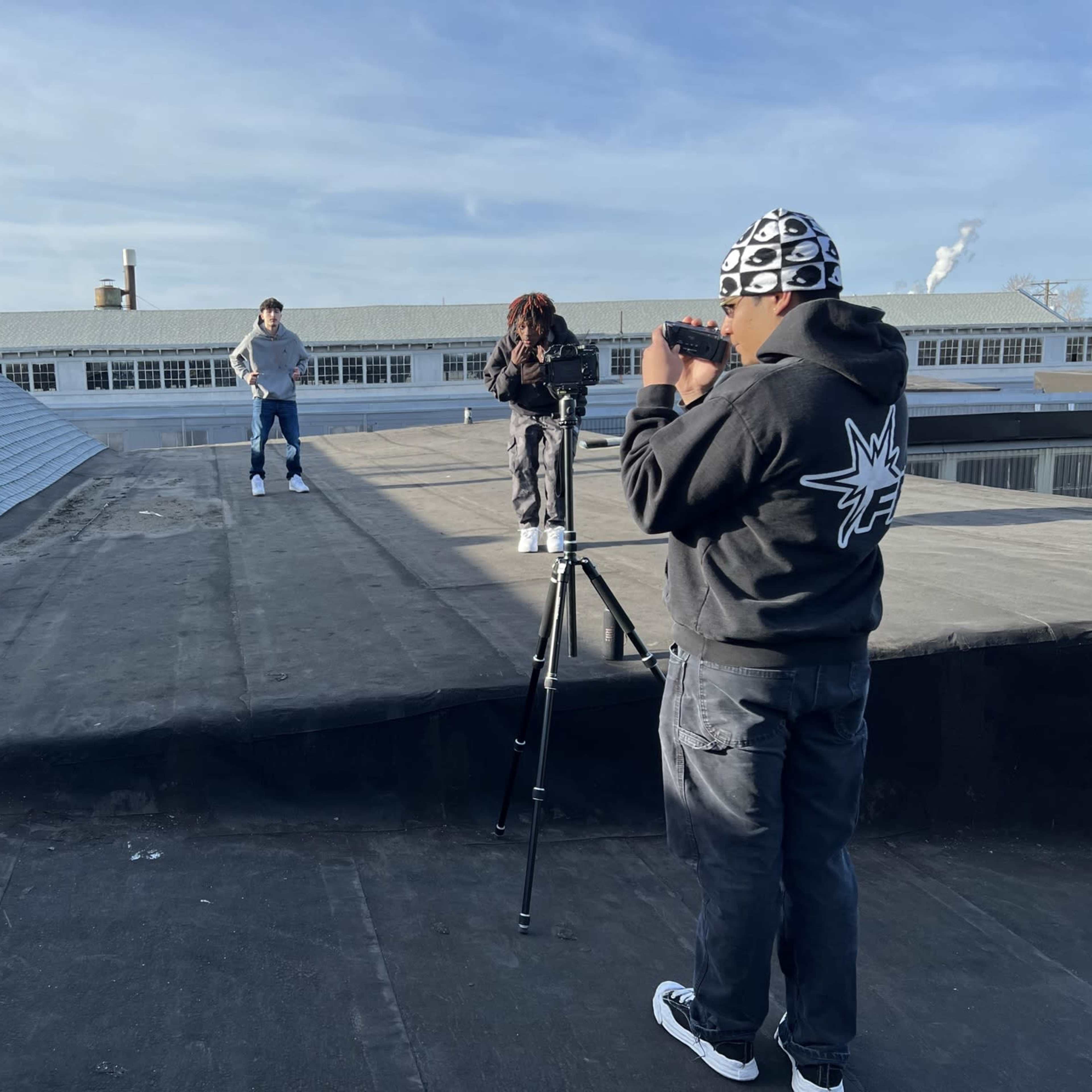 Three individuals are on a rooftop, with one person filming another who is posing in front of a camera.