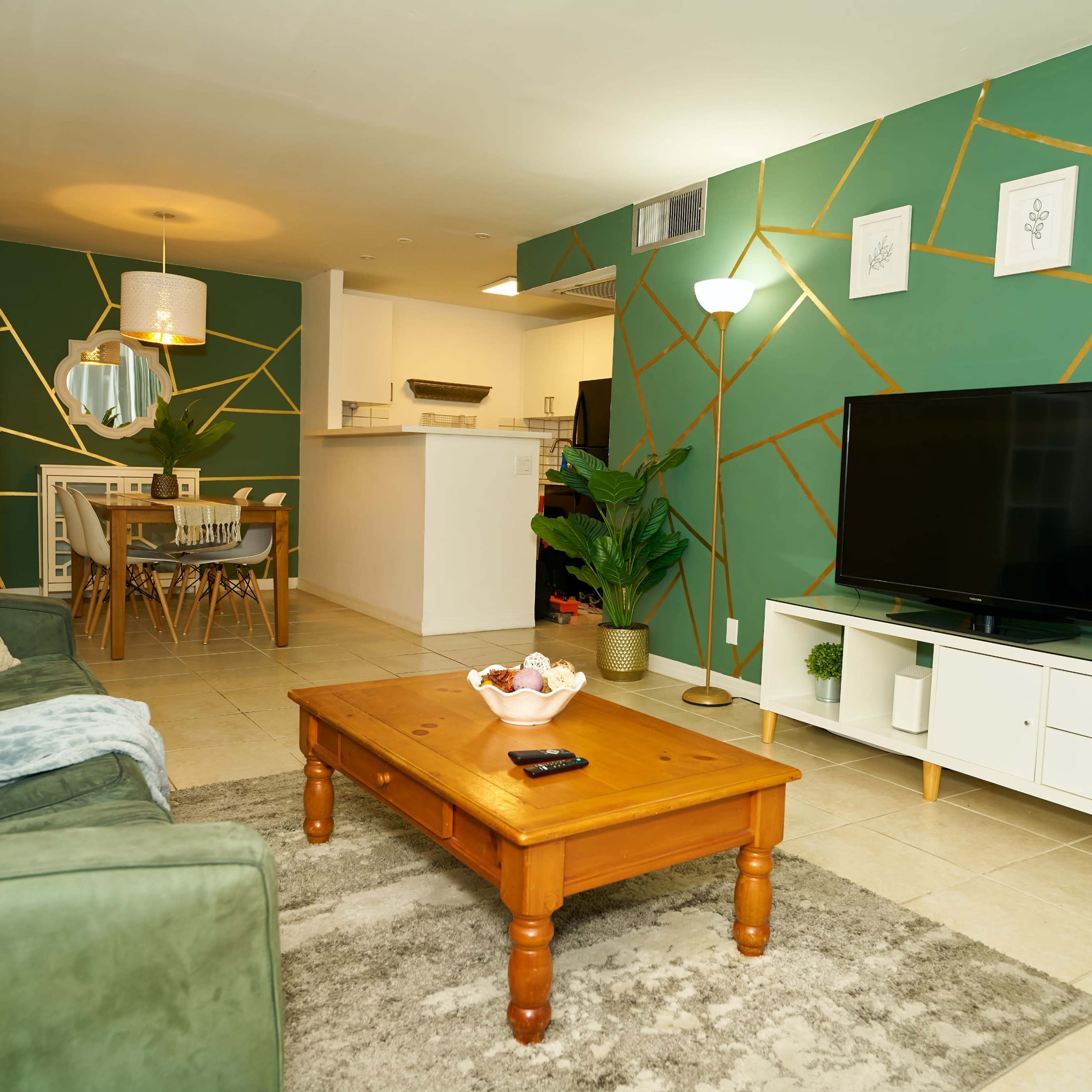 A modern living room features a green accent wall with gold geometric lines, a wooden coffee table, a television on a white stand, and a dining area visible in the background.