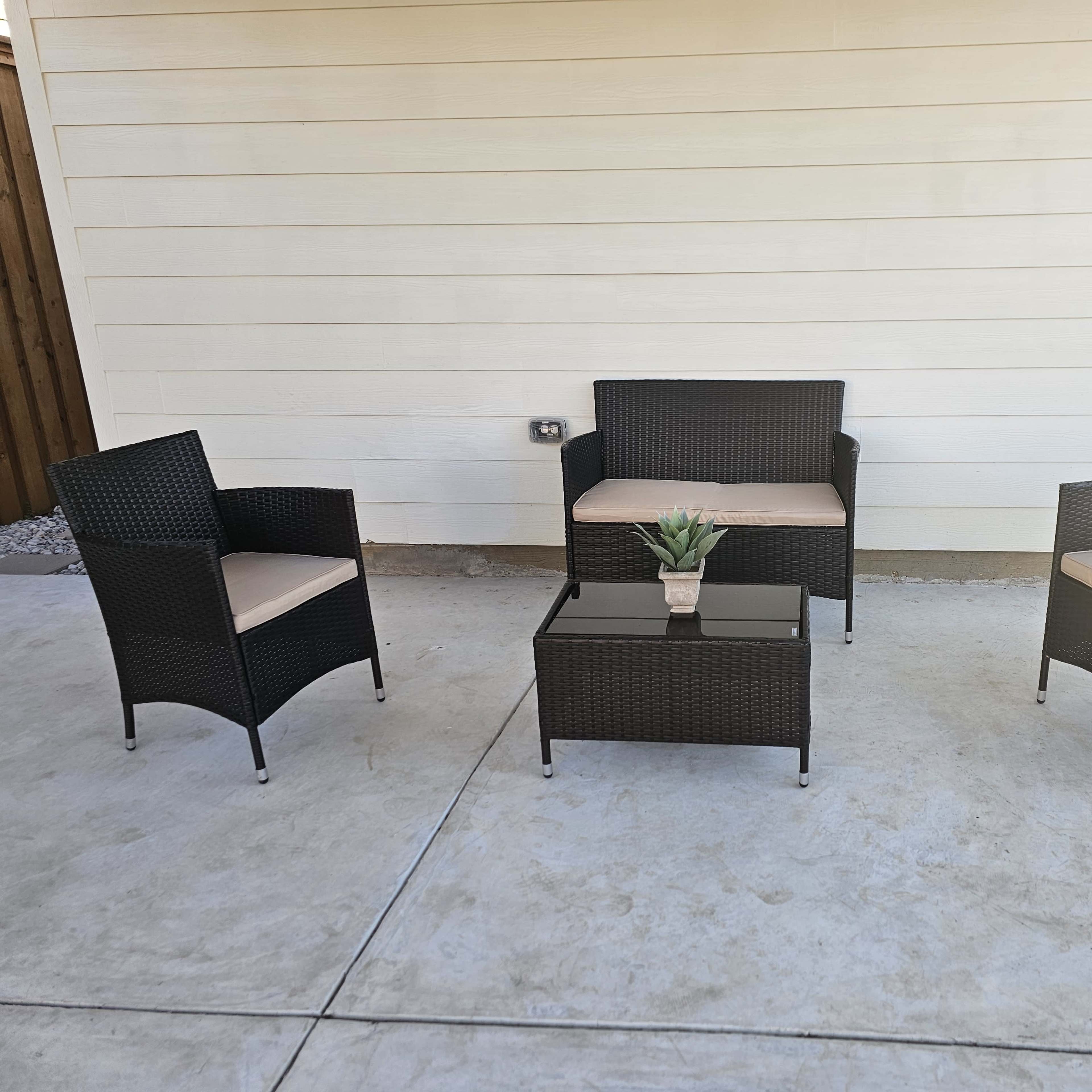A small outdoor seating area features two armchairs and a loveseat around a glass-topped table, all made of black wicker with cream-colored cushions.