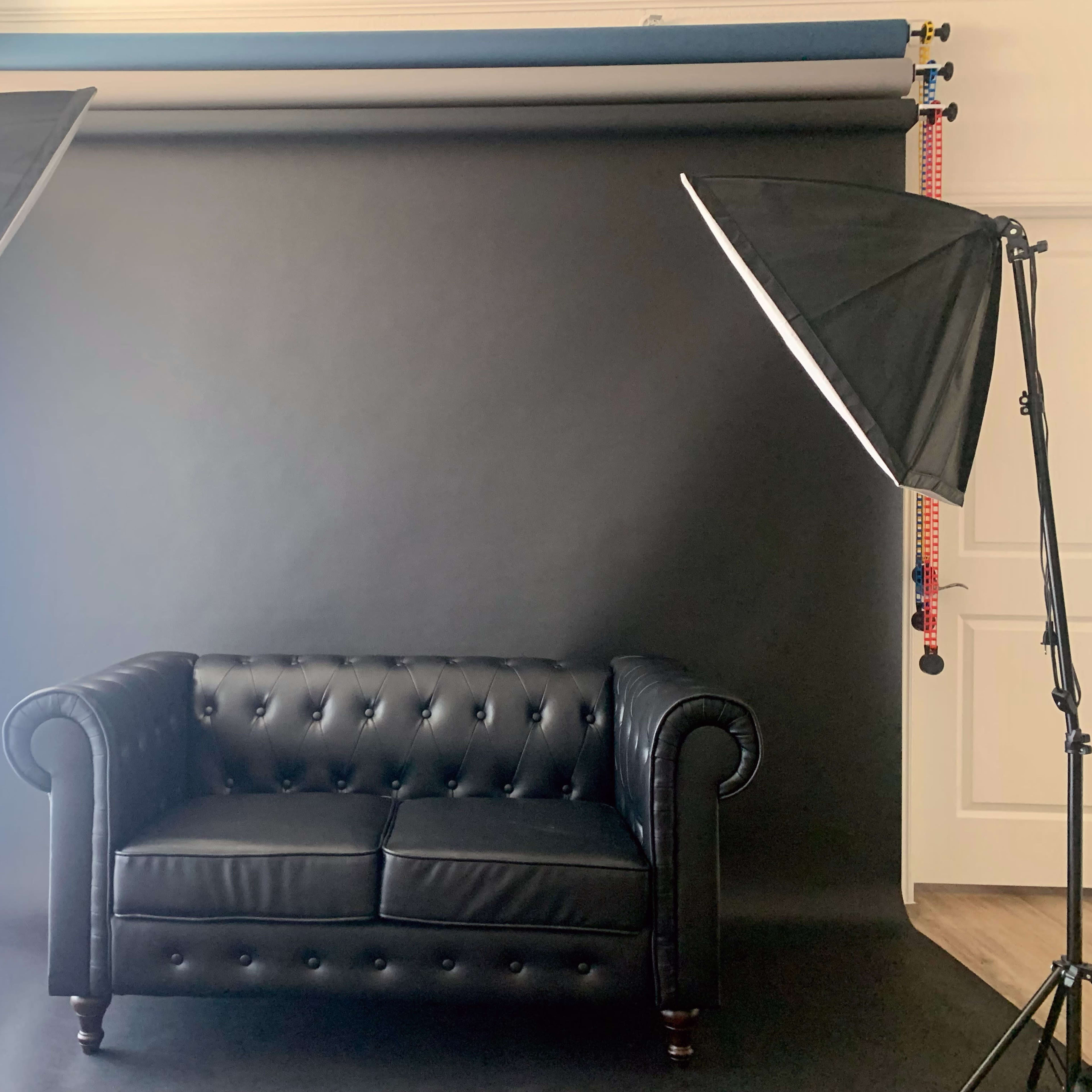 A black leather chesterfield sofa is positioned in front of a dark backdrop, illuminated by two softbox lights.