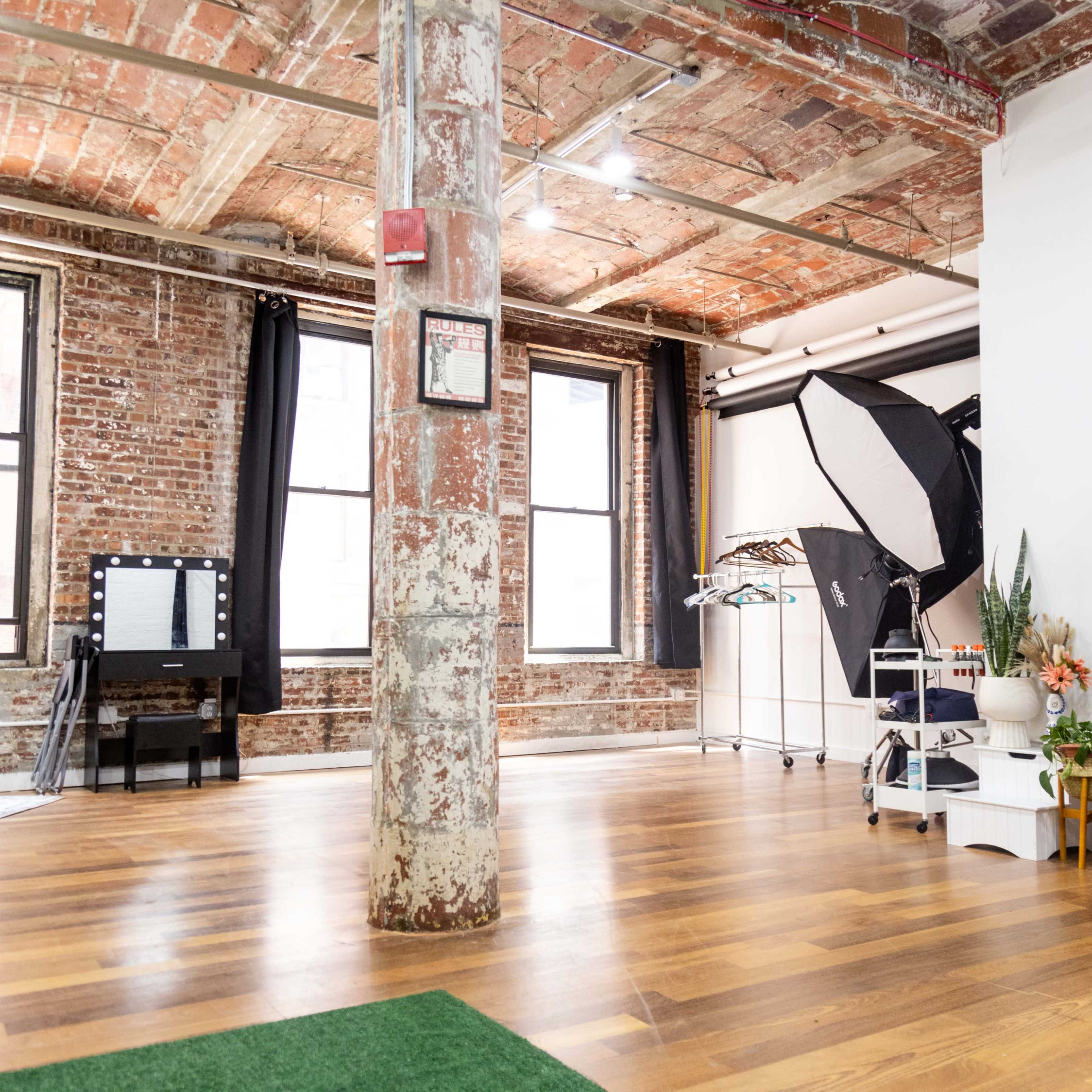 An industrial-style room with exposed brick walls, wooden flooring, and various photography equipment, including a reflector and a rack of clothes.