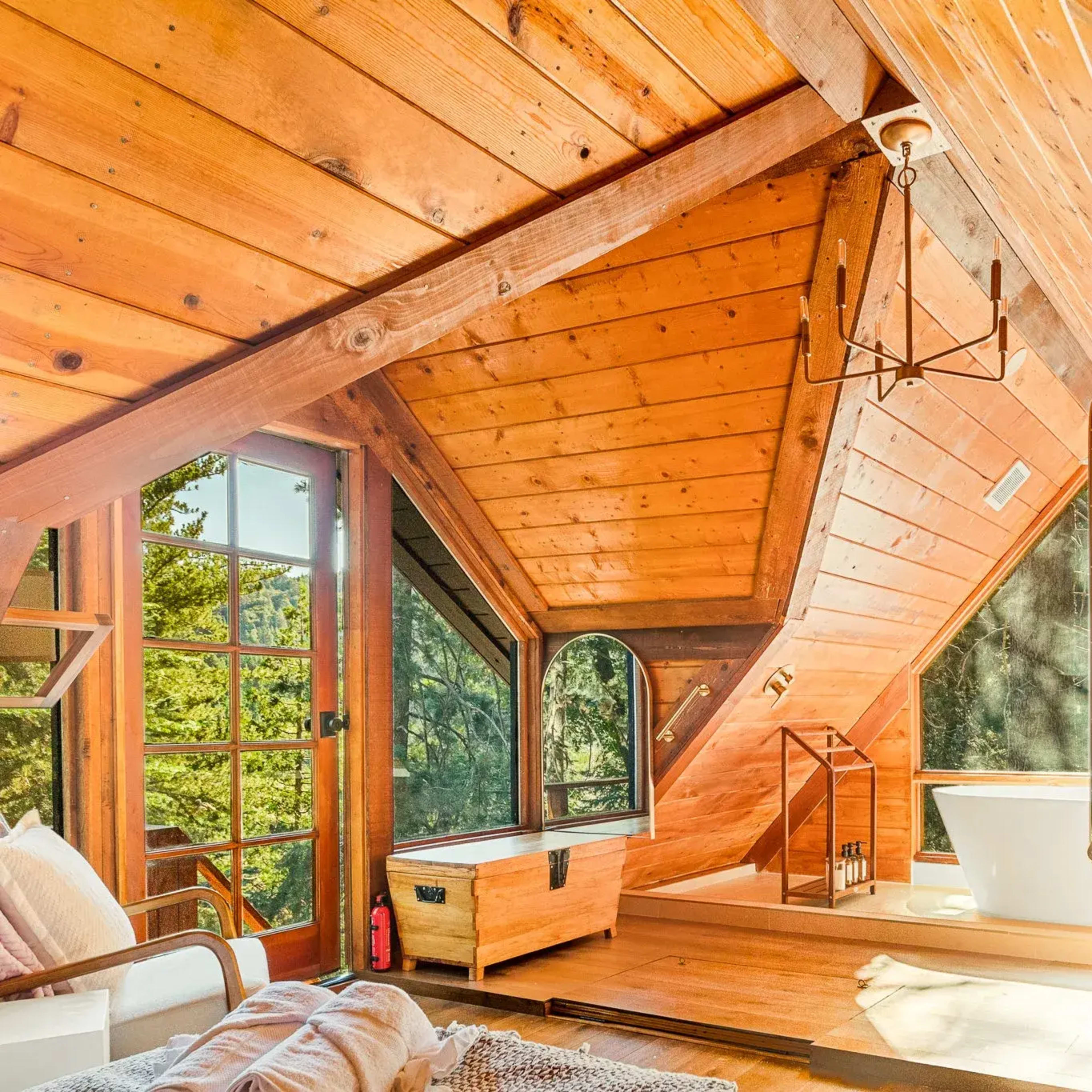 A wooden interior of an A-frame cabin featuring large windows, a cozy seating area, and a freestanding bathtub illuminated by natural light.