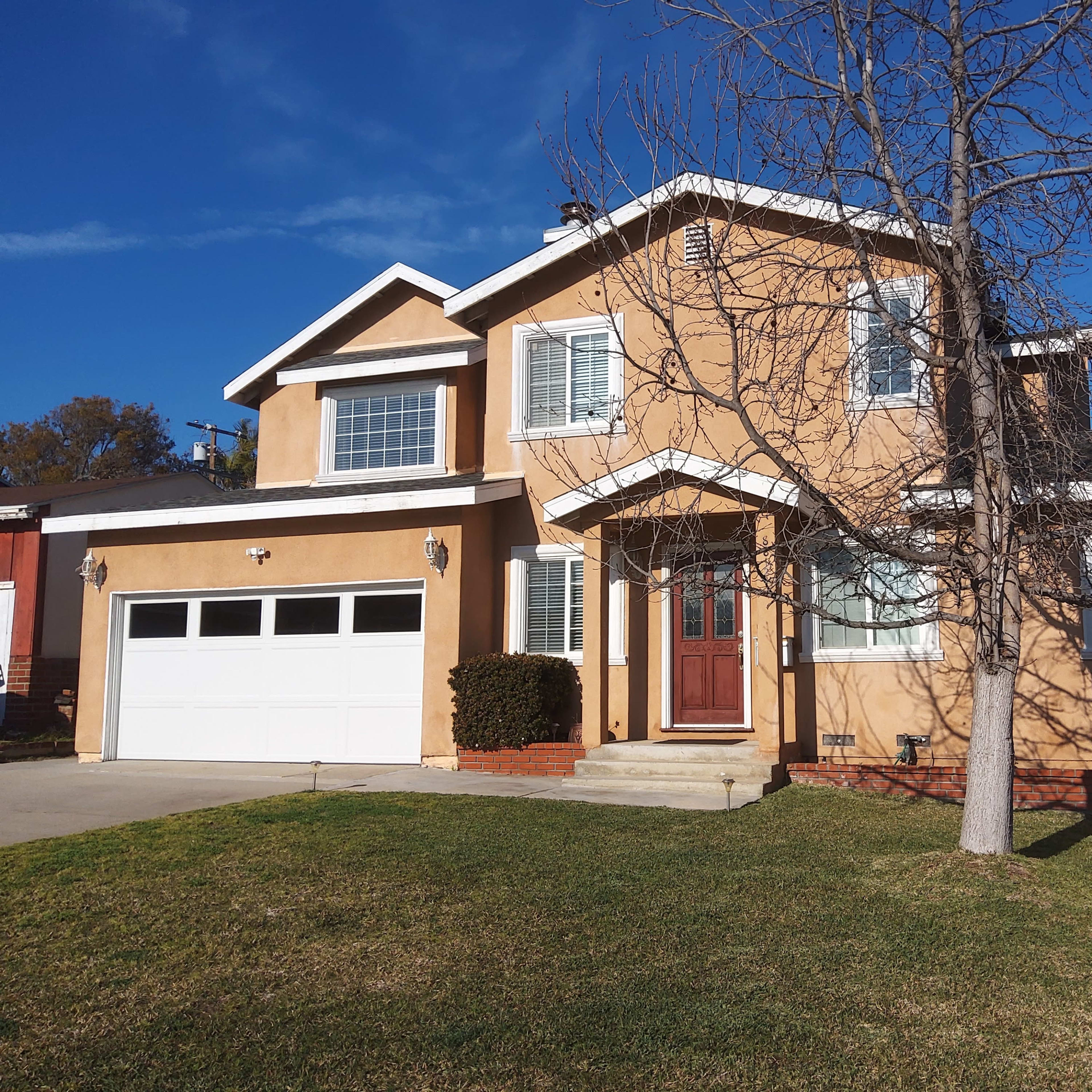 A two-story, tan-colored house with a front porch, a large garage, and a tree in the yard.