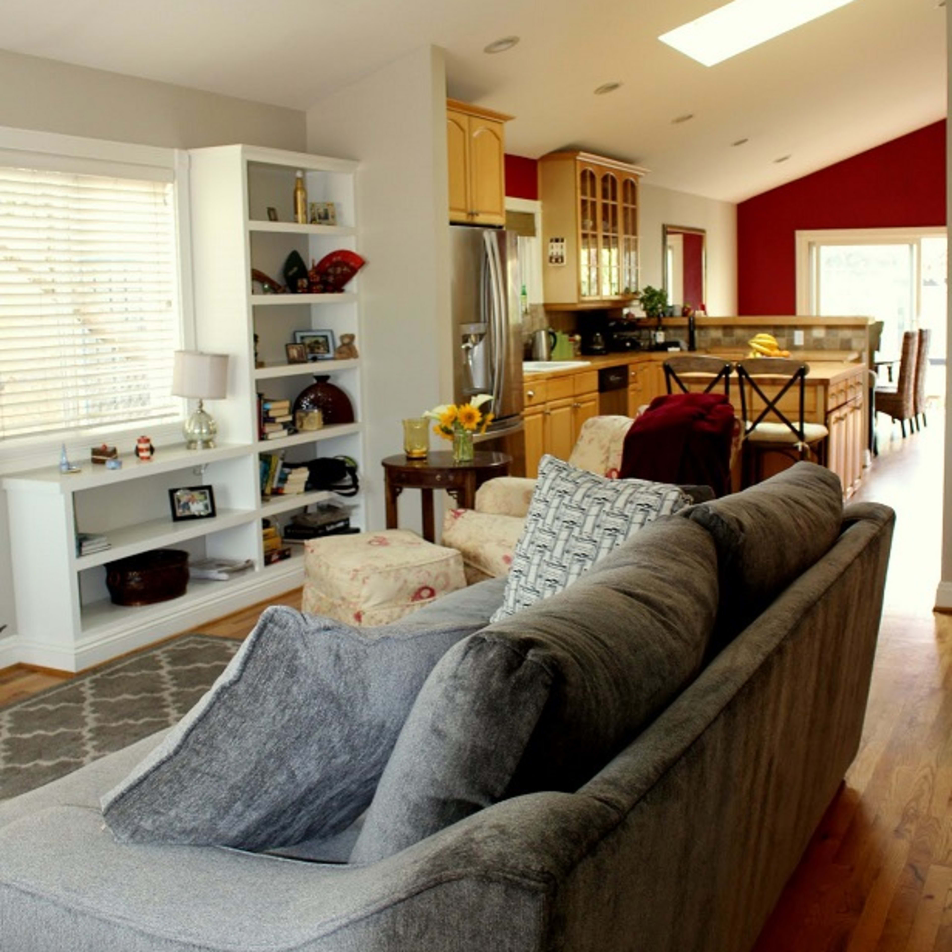 A spacious living area that opens into a kitchen, featuring a sectional couch, a dining table in the background, and large windows allowing natural light.