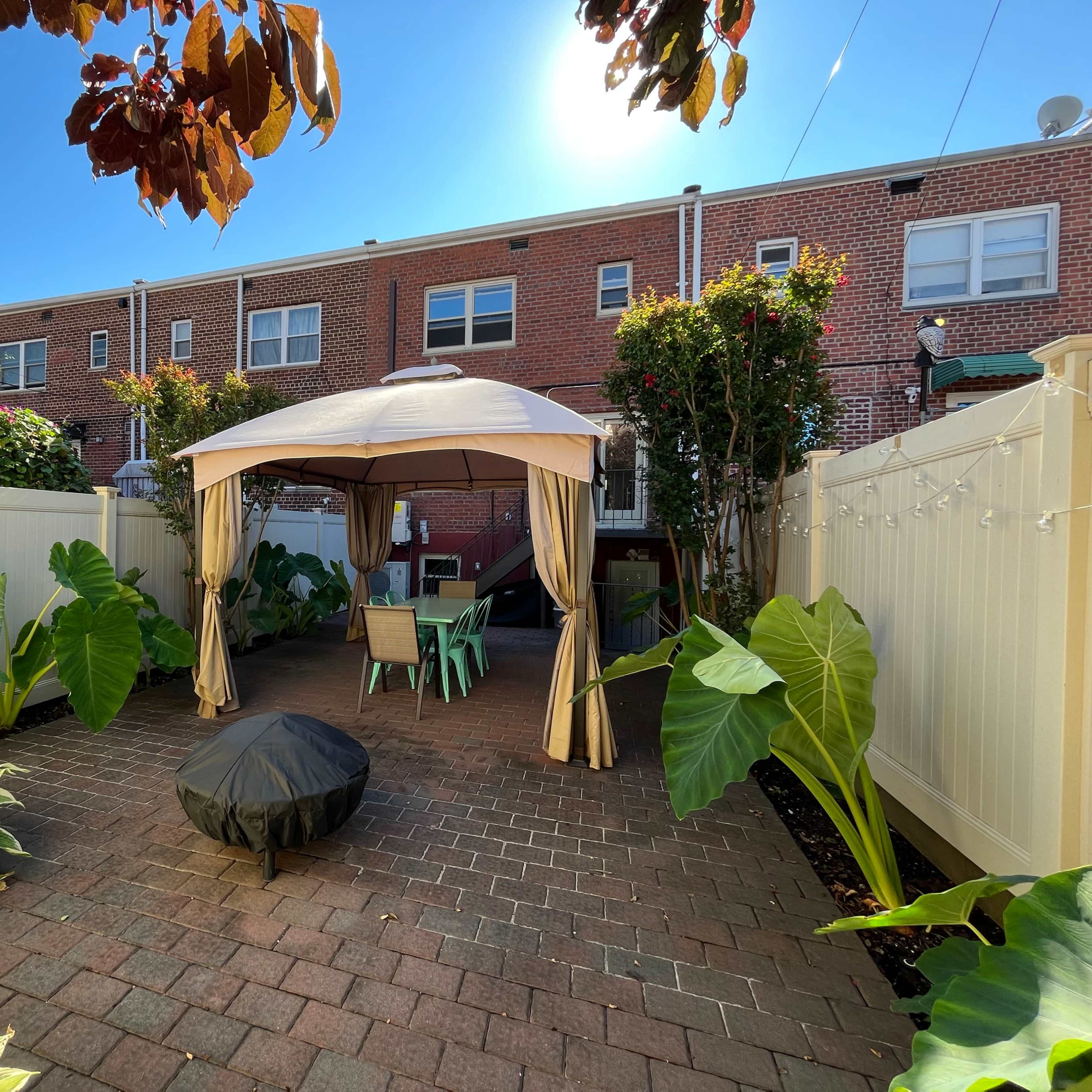 A brick patio features a gazebo surrounded by large foliage and outdoor seating in a residential backyard.