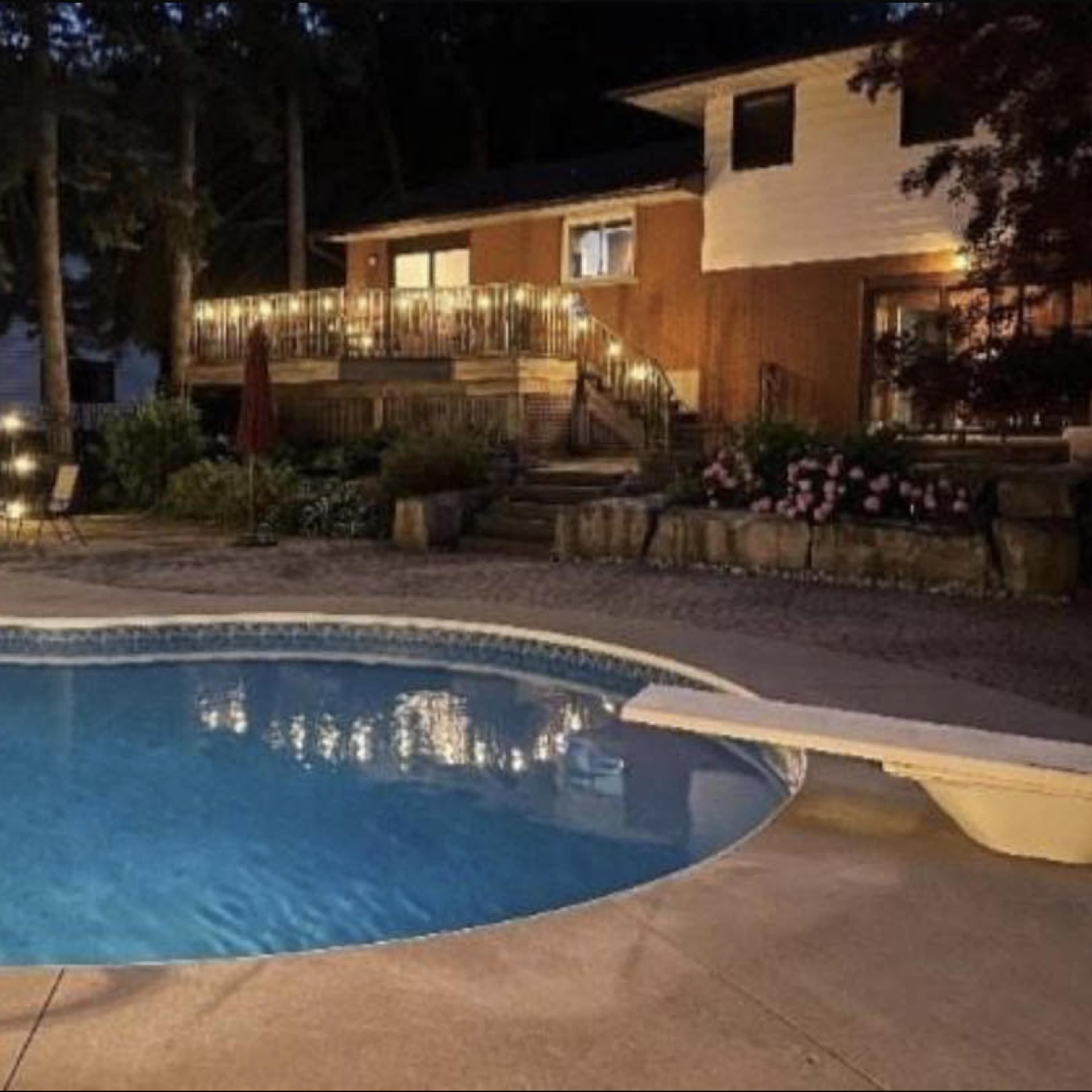 A well-lit backyard pool area at dusk, featuring a pool with a diving board, surrounded by trees and a house with outdoor lighting.