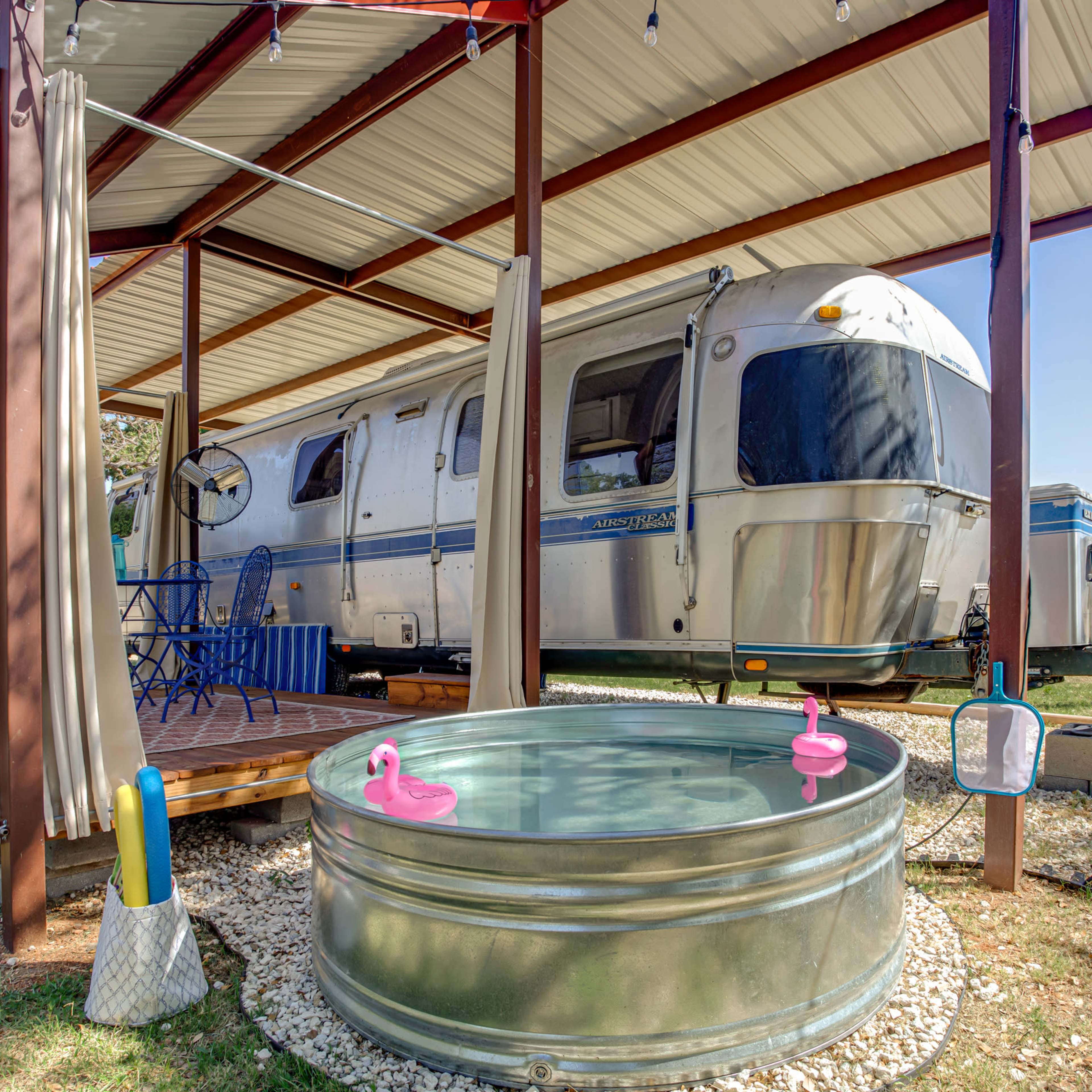 An Airstream trailer is parked under a metal canopy with a wooden deck and a small galvanized tub in front, adorned with pink rubber ducks.