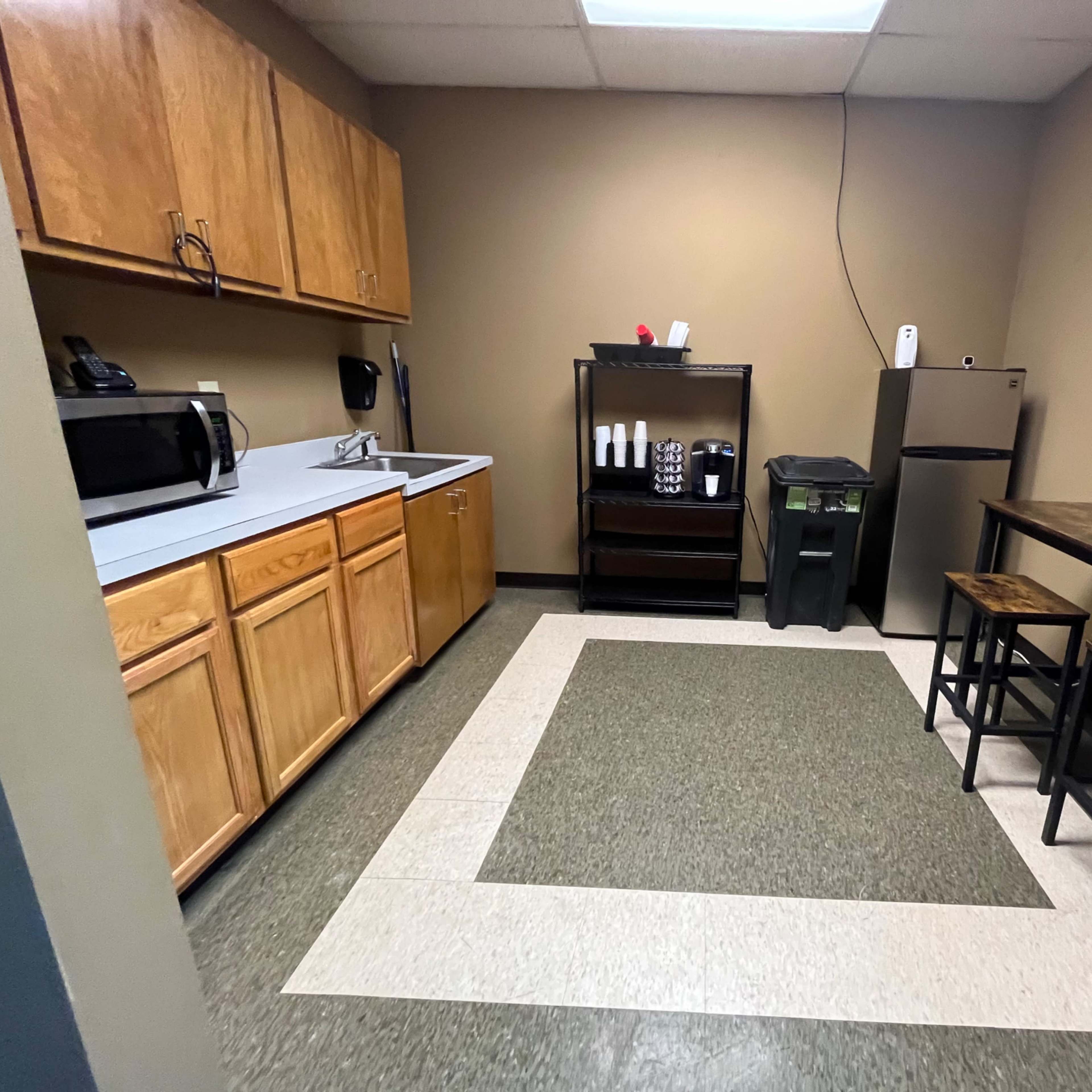 A small kitchen area featuring wooden cabinets, a sink, a microwave, a coffee station, a trash can, and a table with two stools.