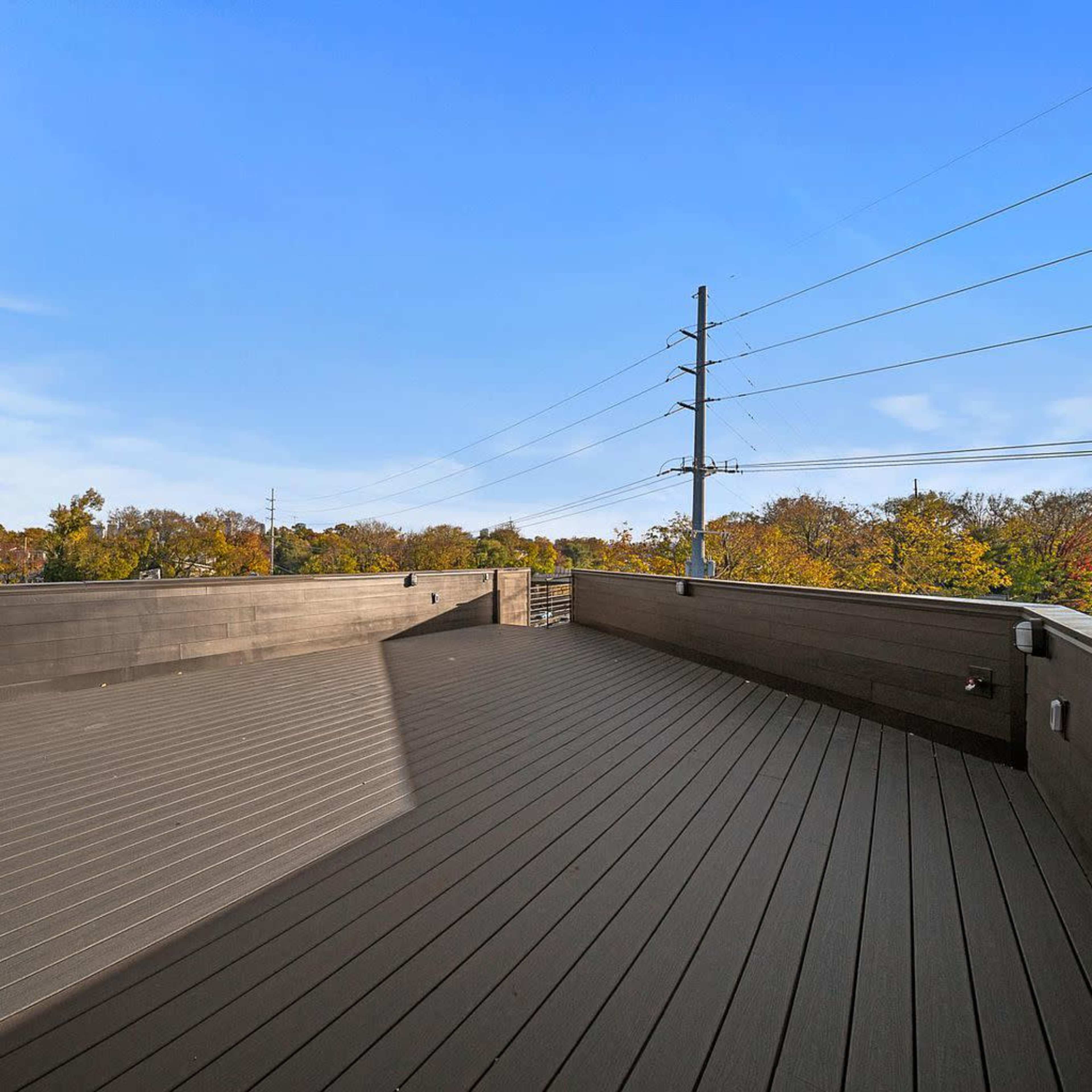 A spacious rooftop deck with wooden flooring overlooks a landscape of trees and power lines under a clear blue sky.