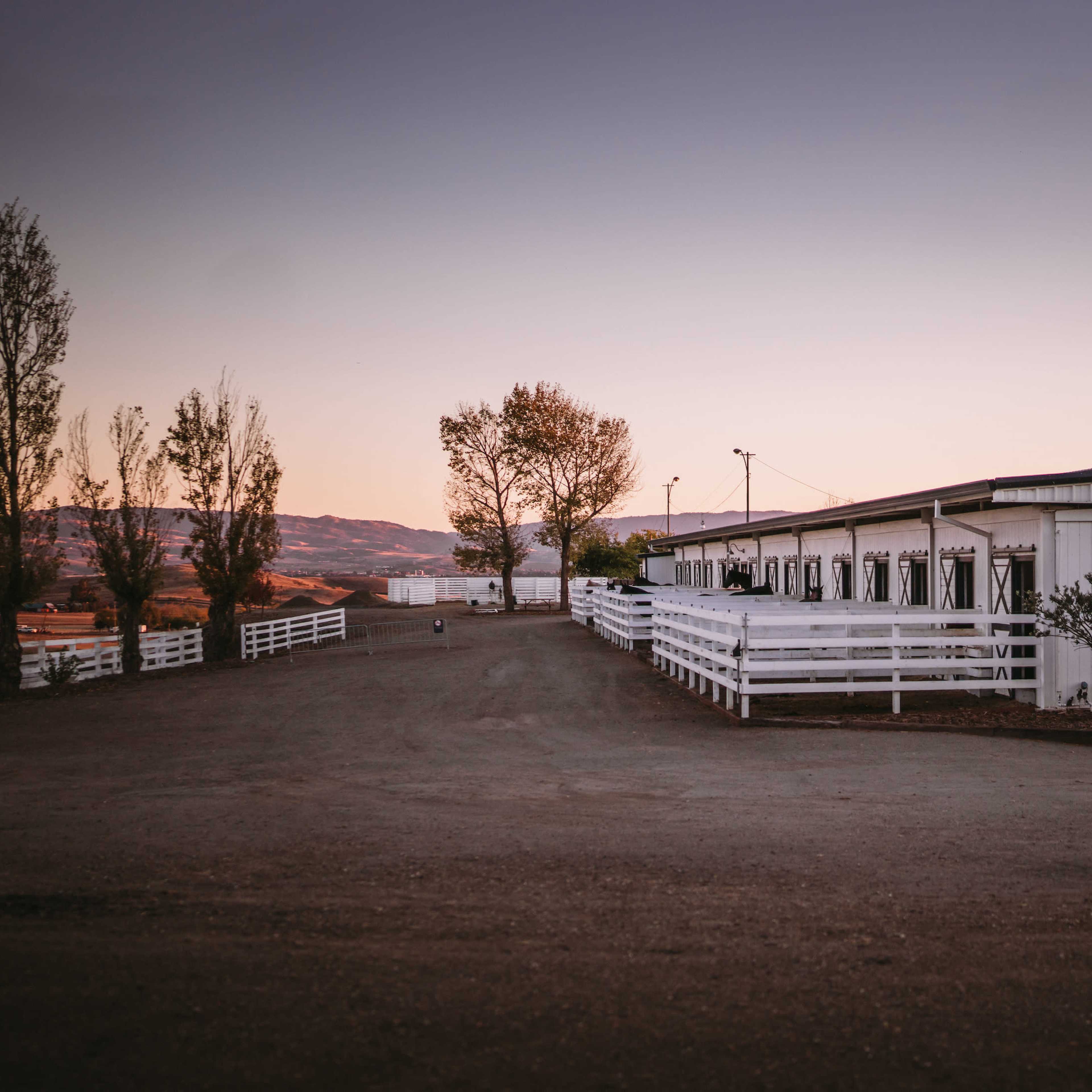A gravel path leads to a white barn with wooden doors, surrounded by trees against a sunset sky.