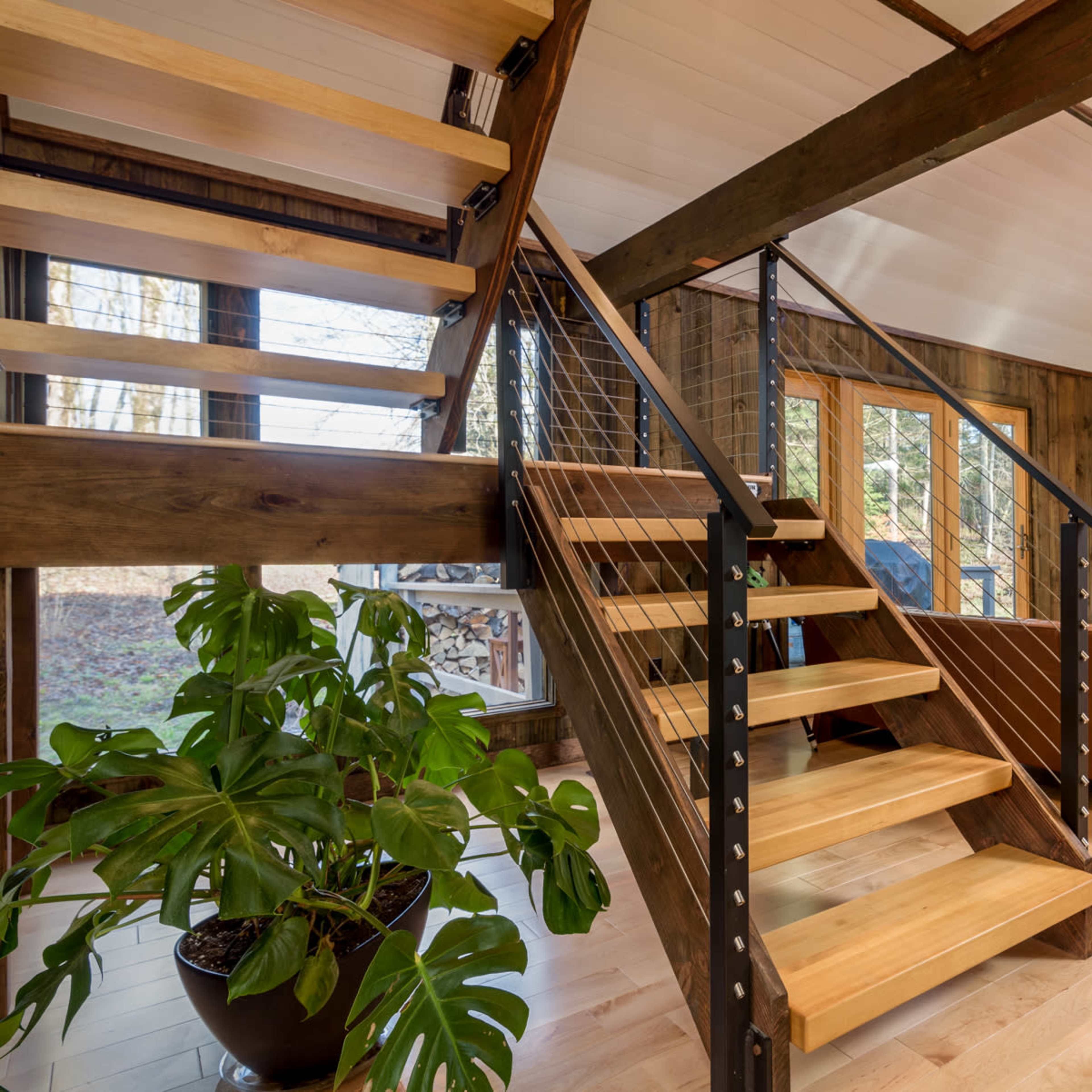 A modern interior with a wooden staircase, a stone fireplace, and large windows overlooking a wooded area.