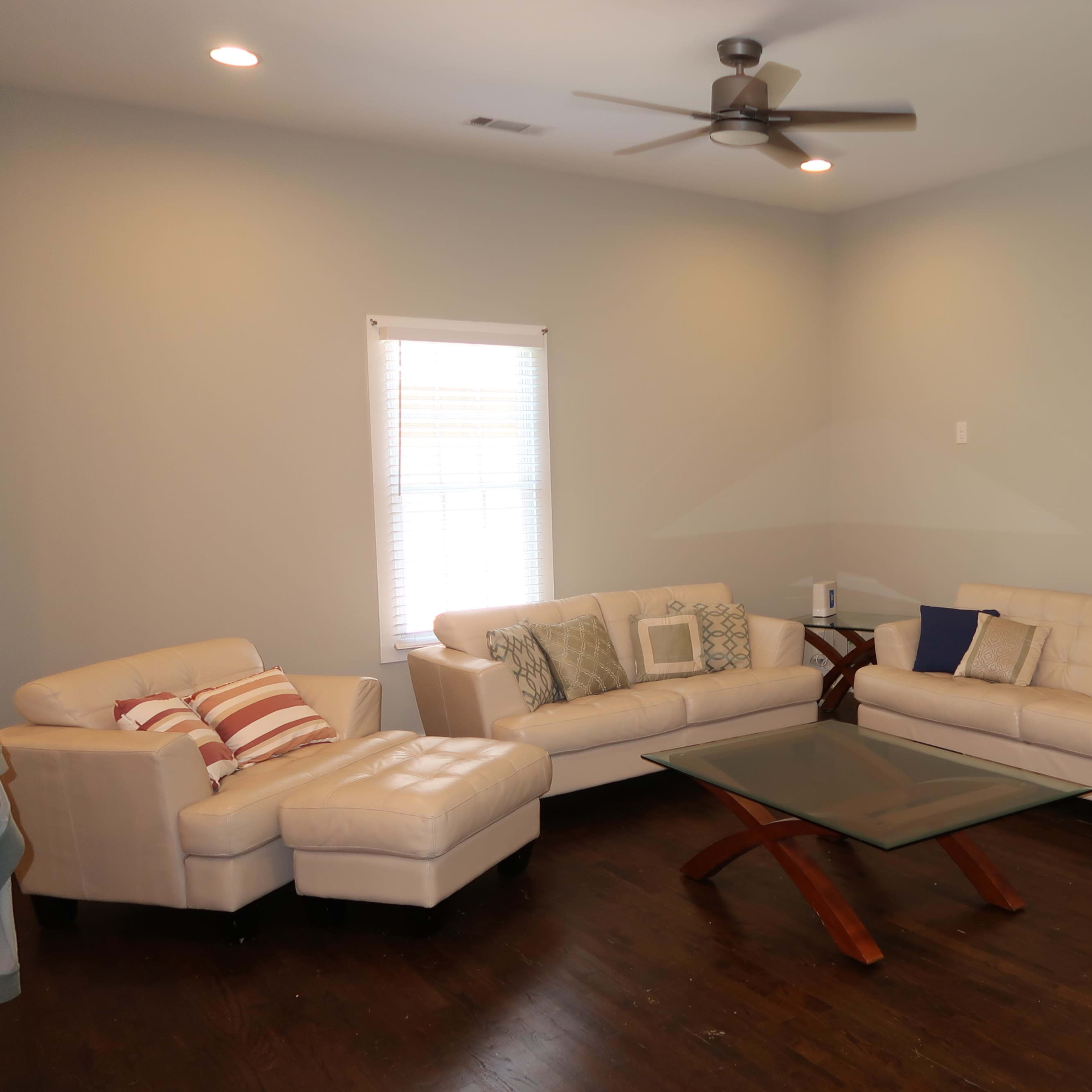 A living room with two light-colored leather sofas, a glass coffee table, a wooden side table with a lamp, and hardwood flooring.