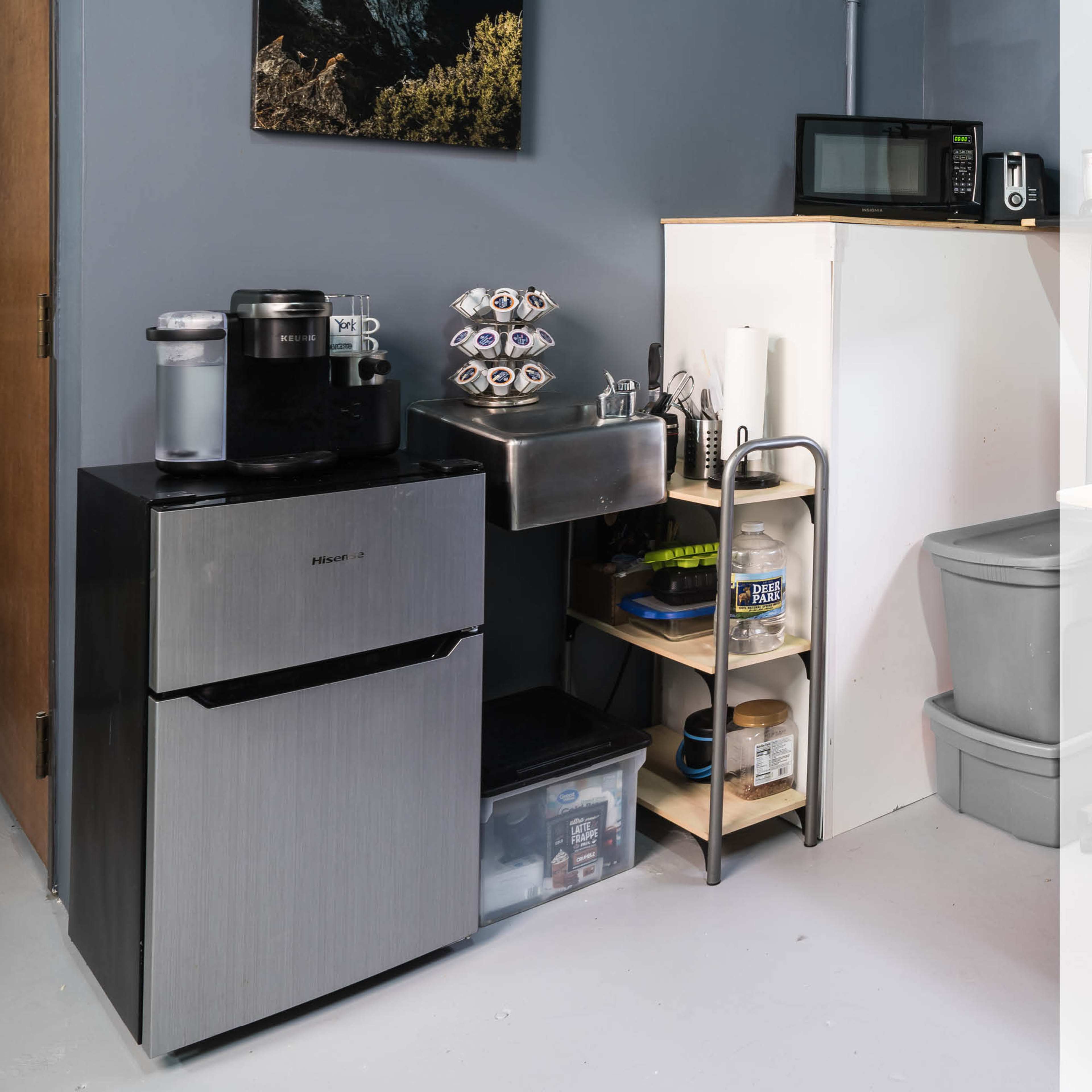 A small kitchenette area with a refrigerator, coffee maker, sink, and shelves stocked with items.