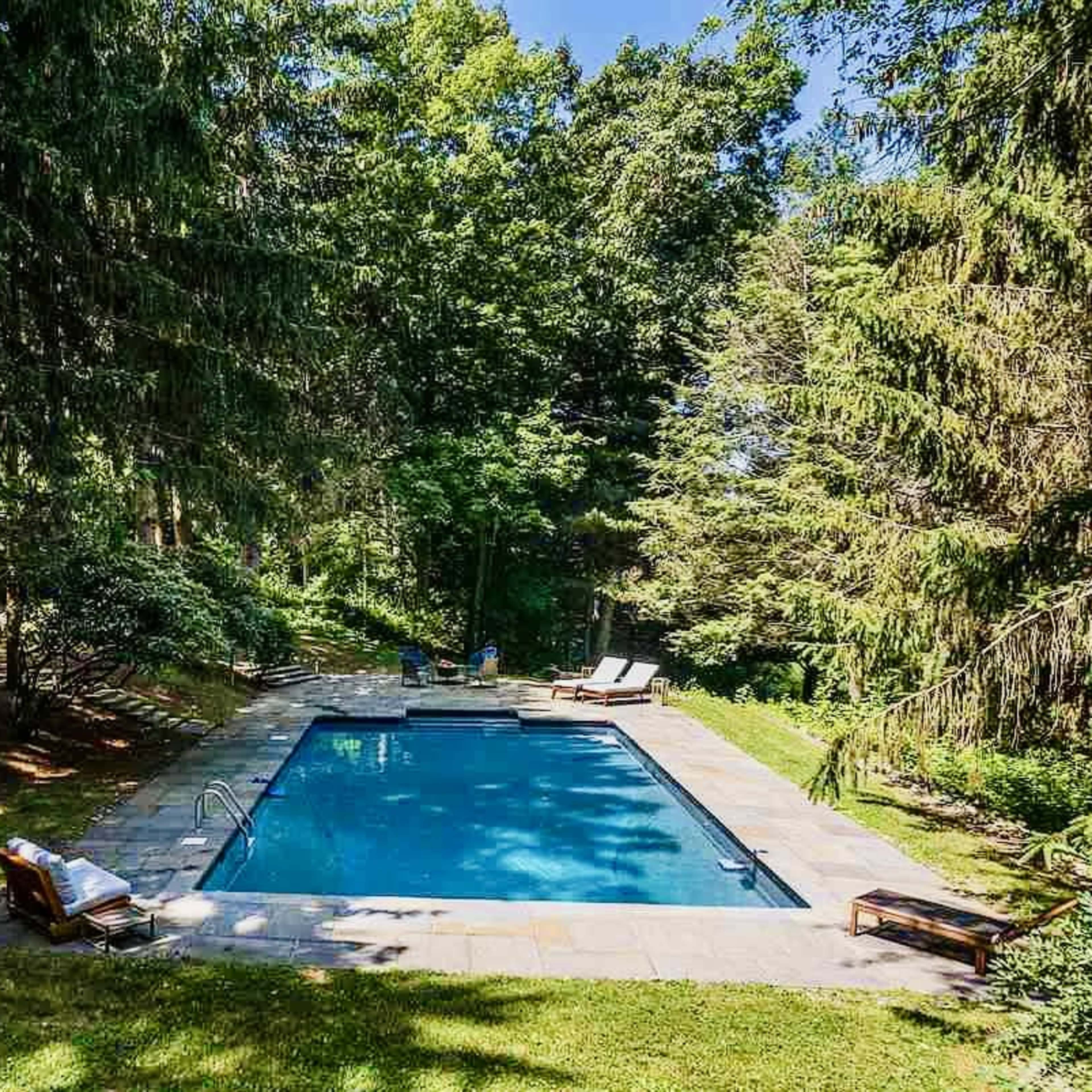 A rectangular swimming pool surrounded by stone paving and lush greenery, with two lounge chairs positioned nearby under the shade of tall trees.