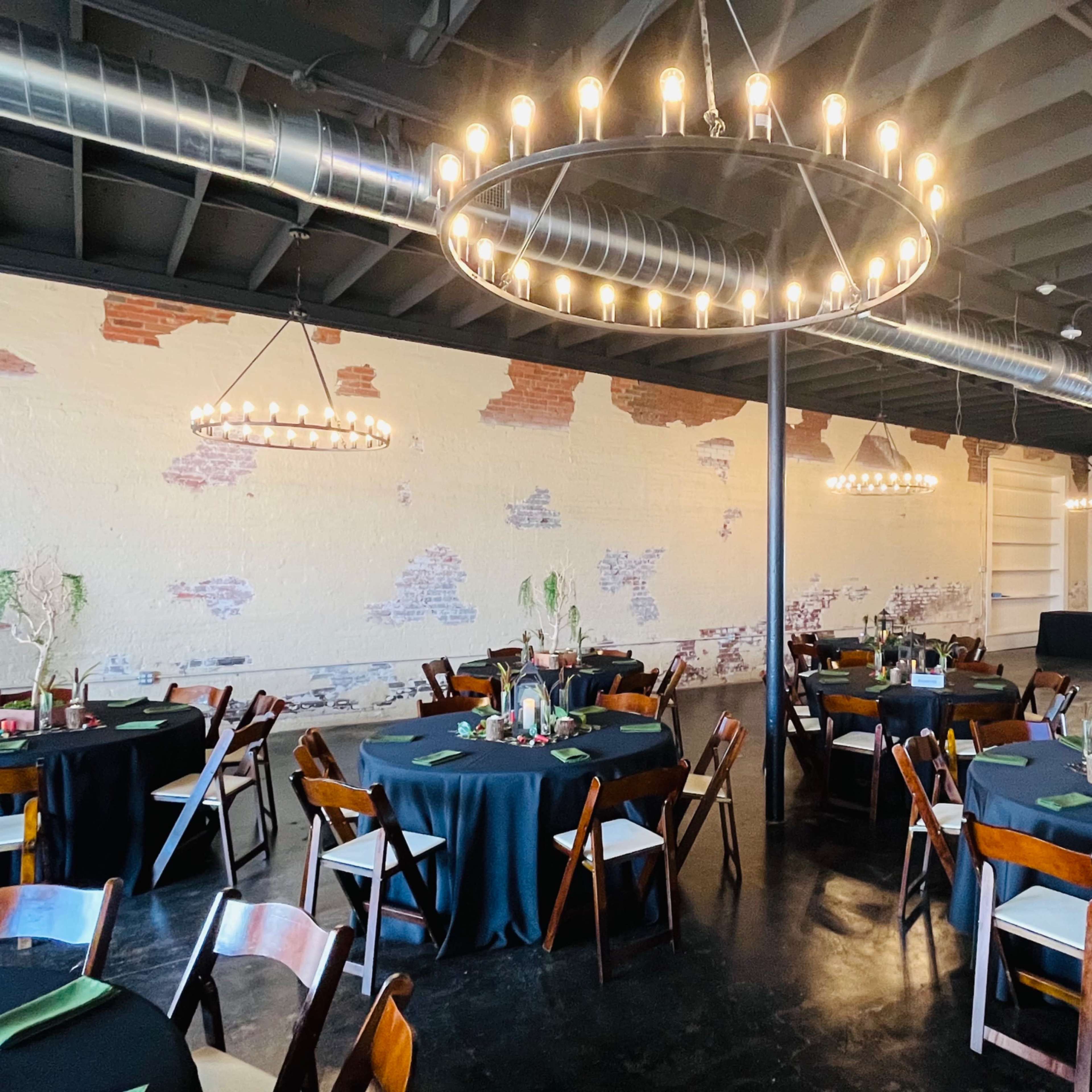 A spacious event venue with round tables covered in black tablecloths, each set with green napkins, and illuminated by large chandeliers hanging from the ceiling.