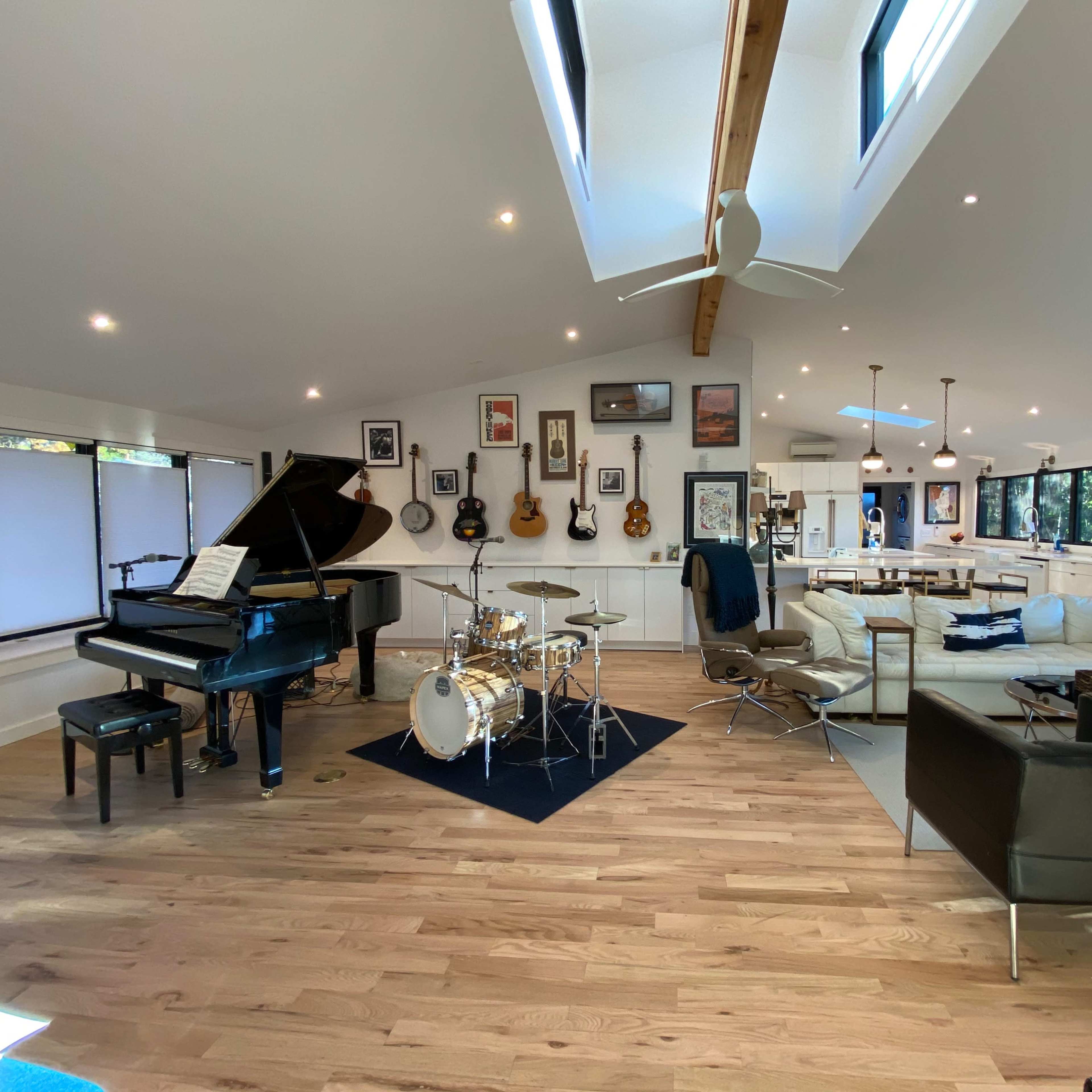 A spacious, well-lit room featuring a grand piano, a drum set, several guitars on the walls, and a mix of seating areas.
