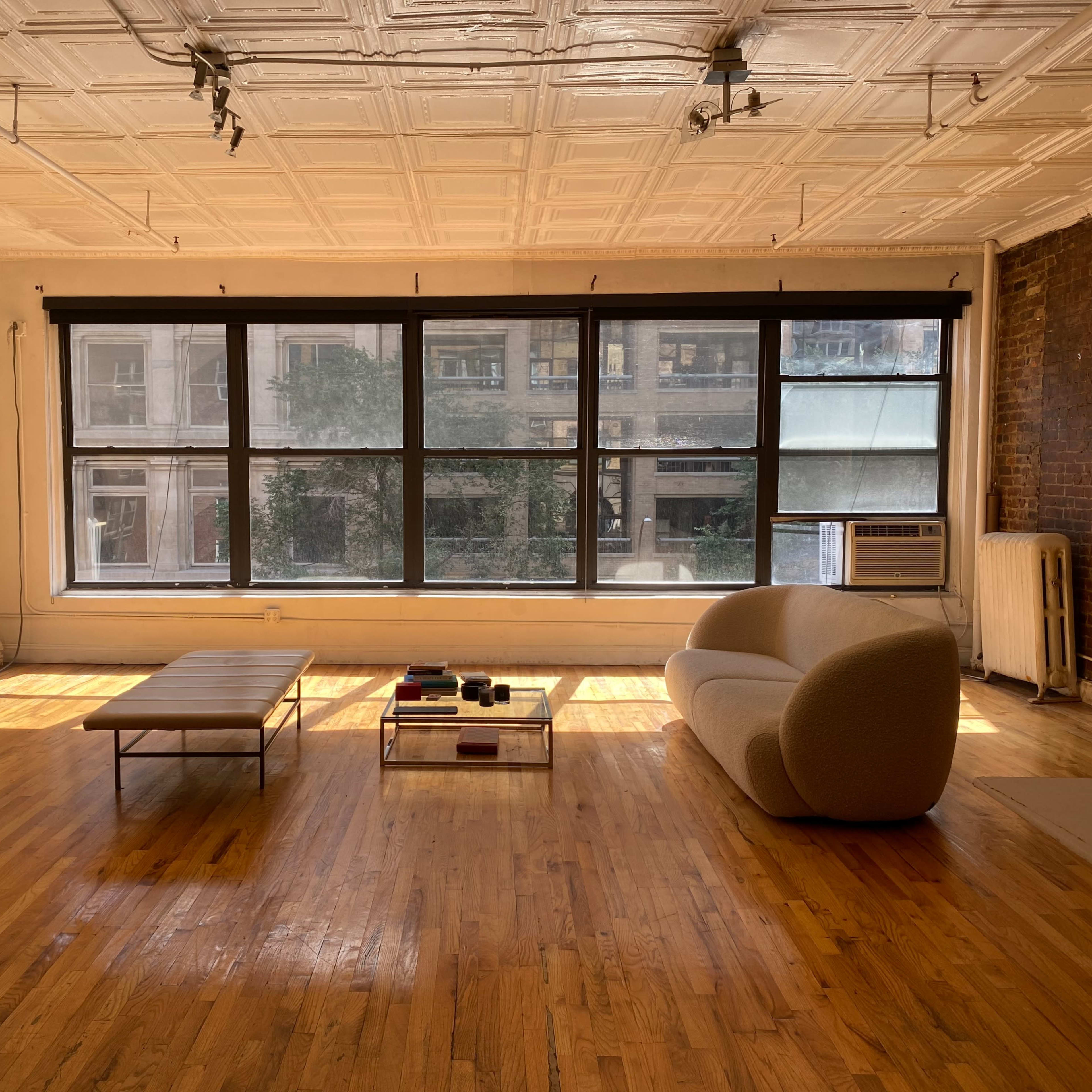 A spacious, sunlit room with large windows, wooden flooring, a simple coffee table, and a light-colored sofa.