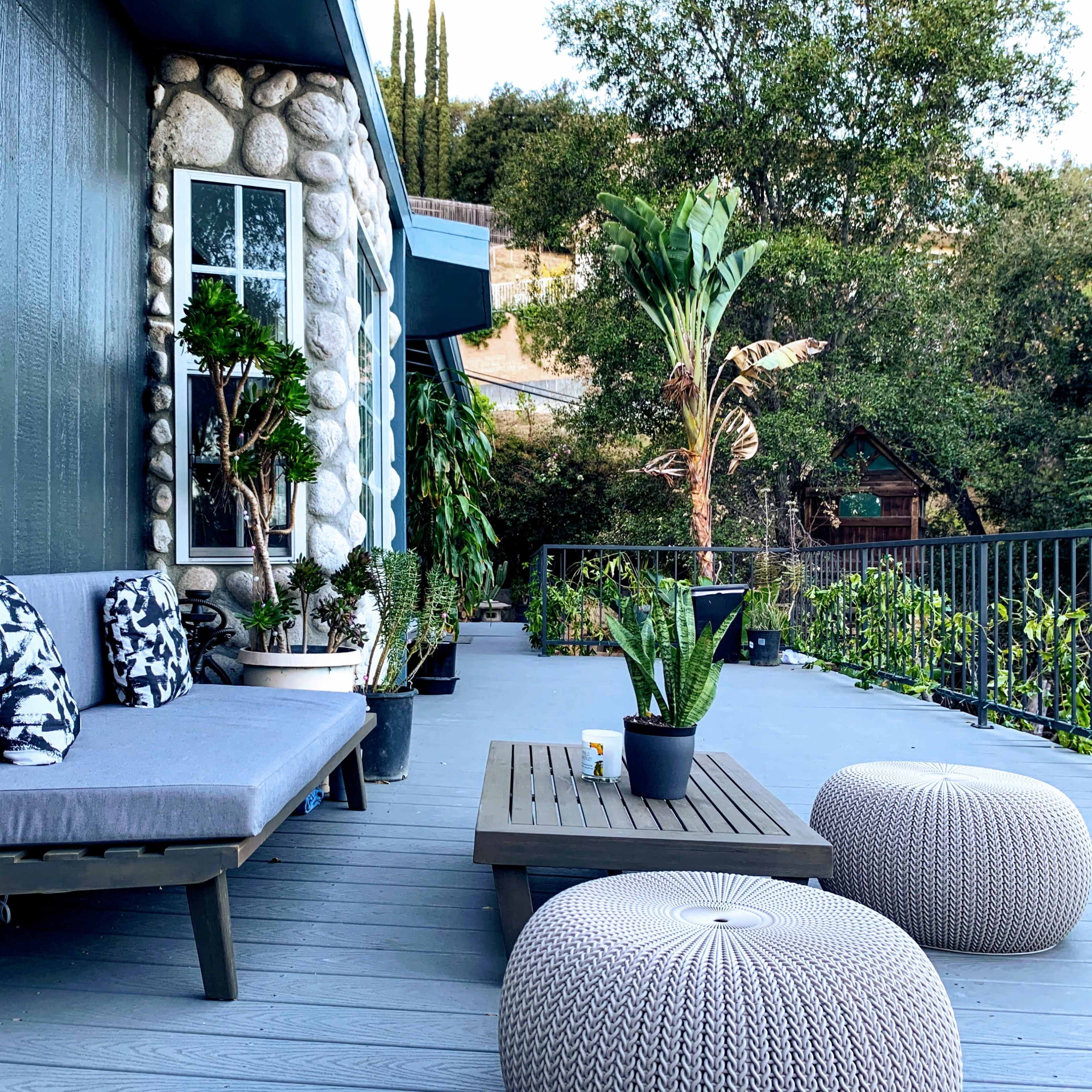A modern patio area with a gray sofa adorned with patterned pillows, surrounded by potted plants and a low wooden coffee table, set against a backdrop of greenery.