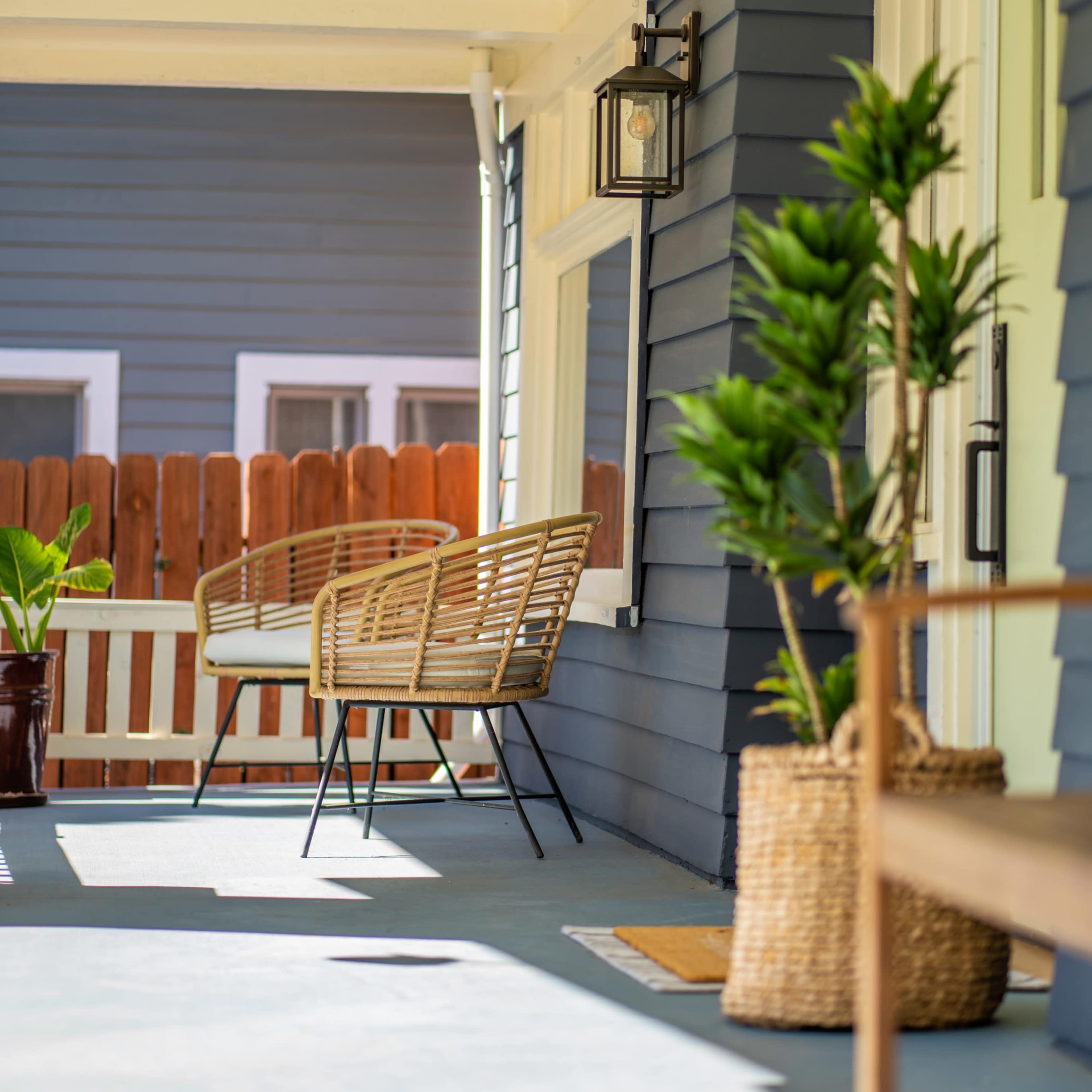 A porch with a rattan bench, potted plants, and a wooden side table against a blue and white house.