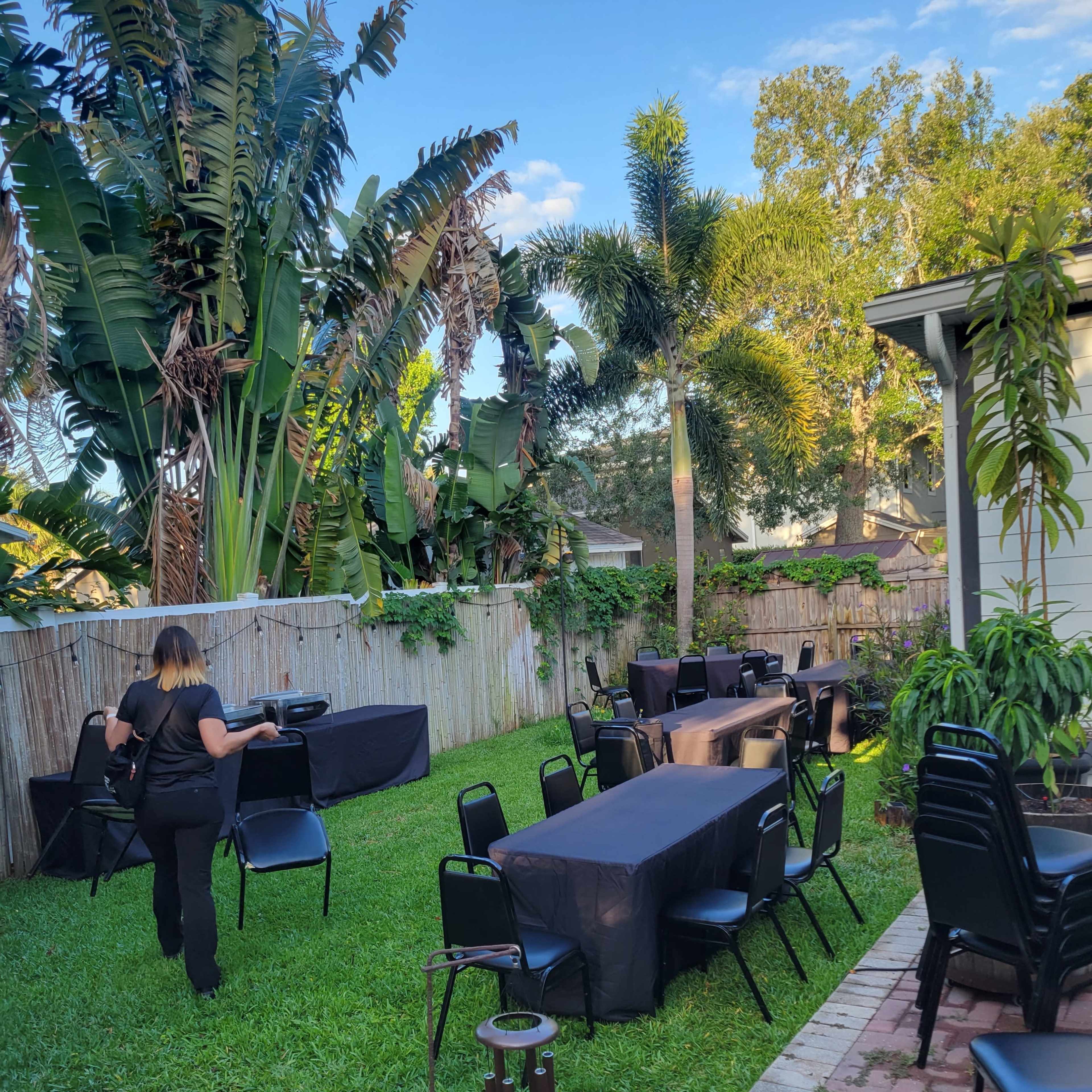 A person sets up black tables and chairs in a grassy backyard surrounded by tropical plants and trees.