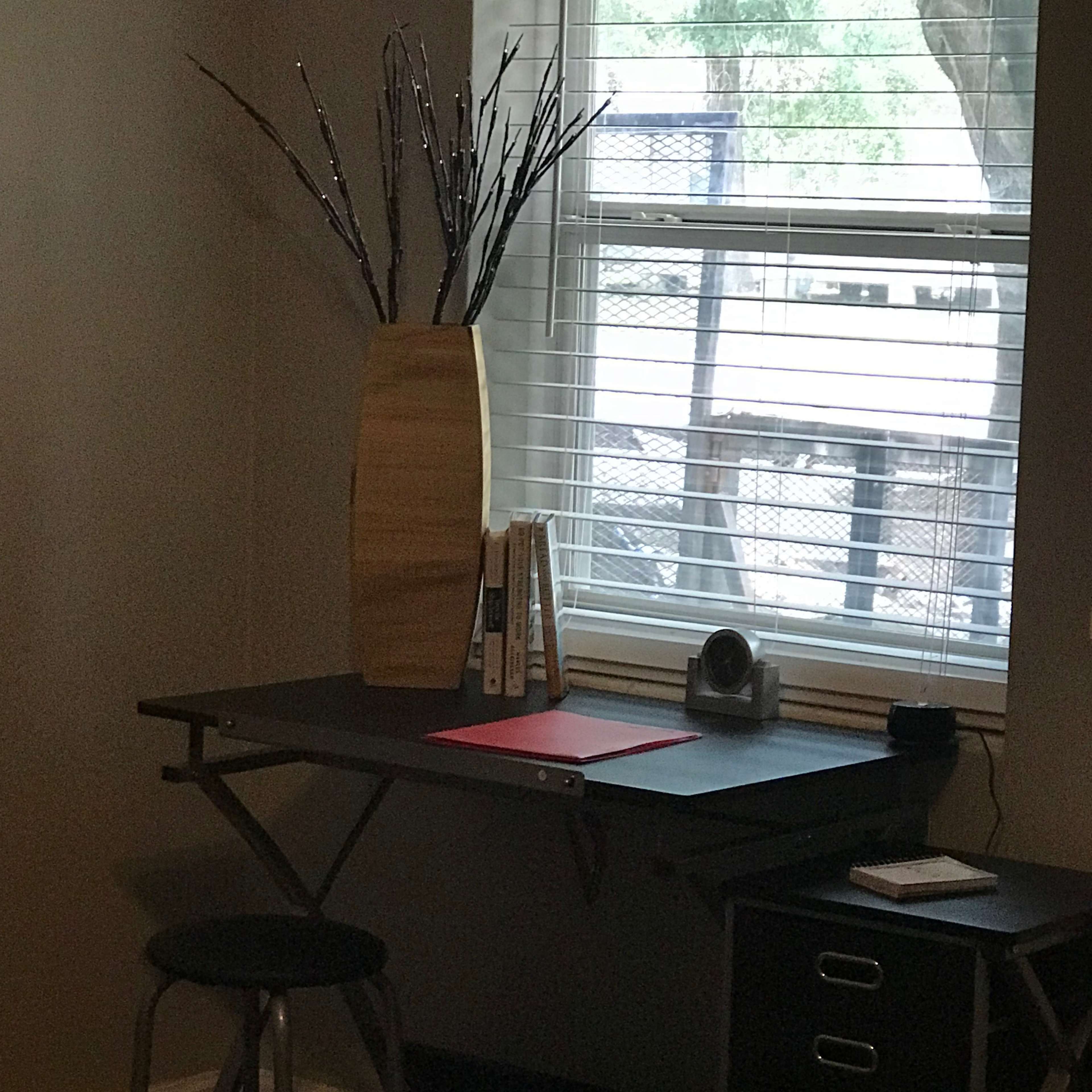 A simple desk with a chair, decorated vase, books, and a camera sits next to a window with blinds.