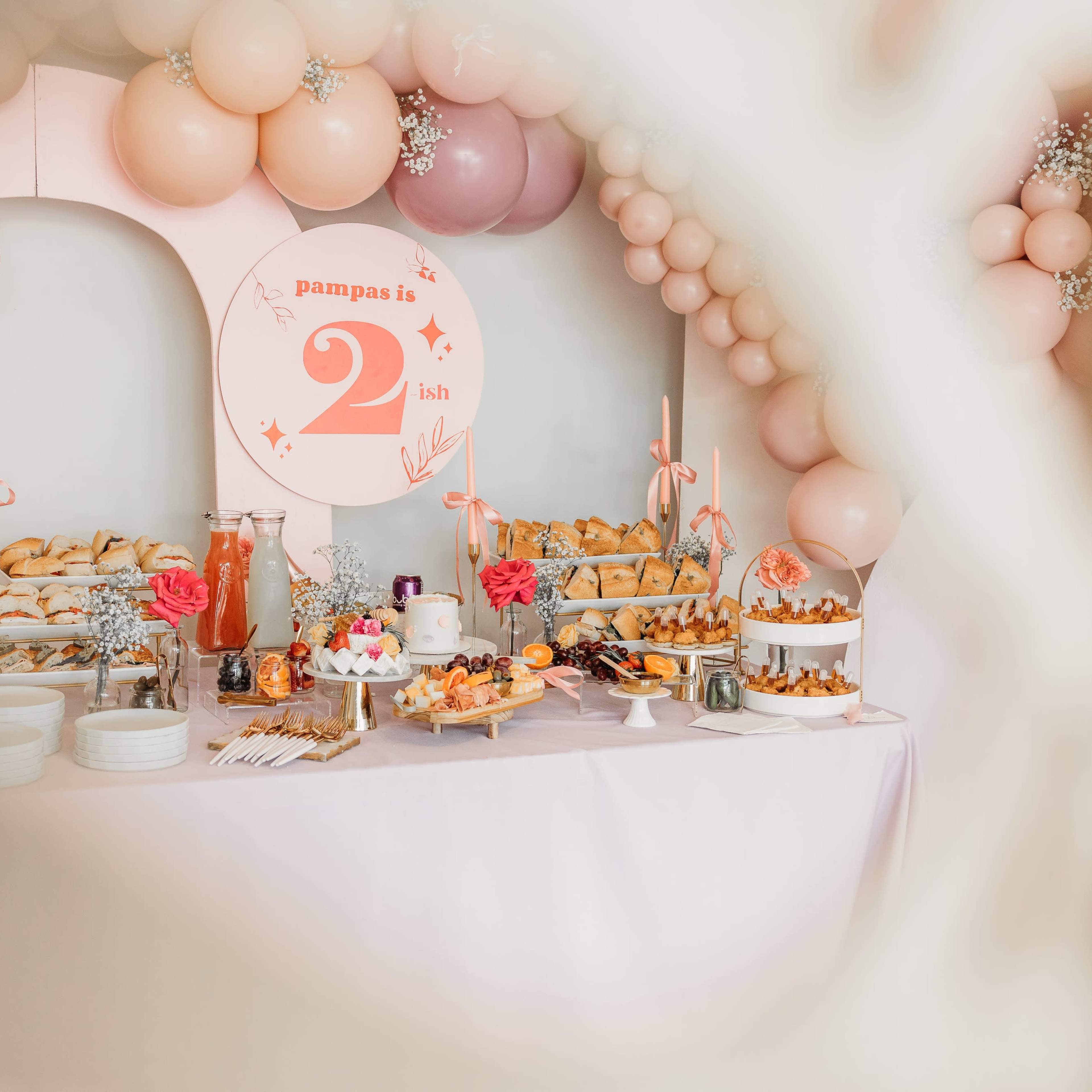 A decorated table is set with various food items, drinks, and a sign celebrating a second birthday, surrounded by pastel-colored balloons.