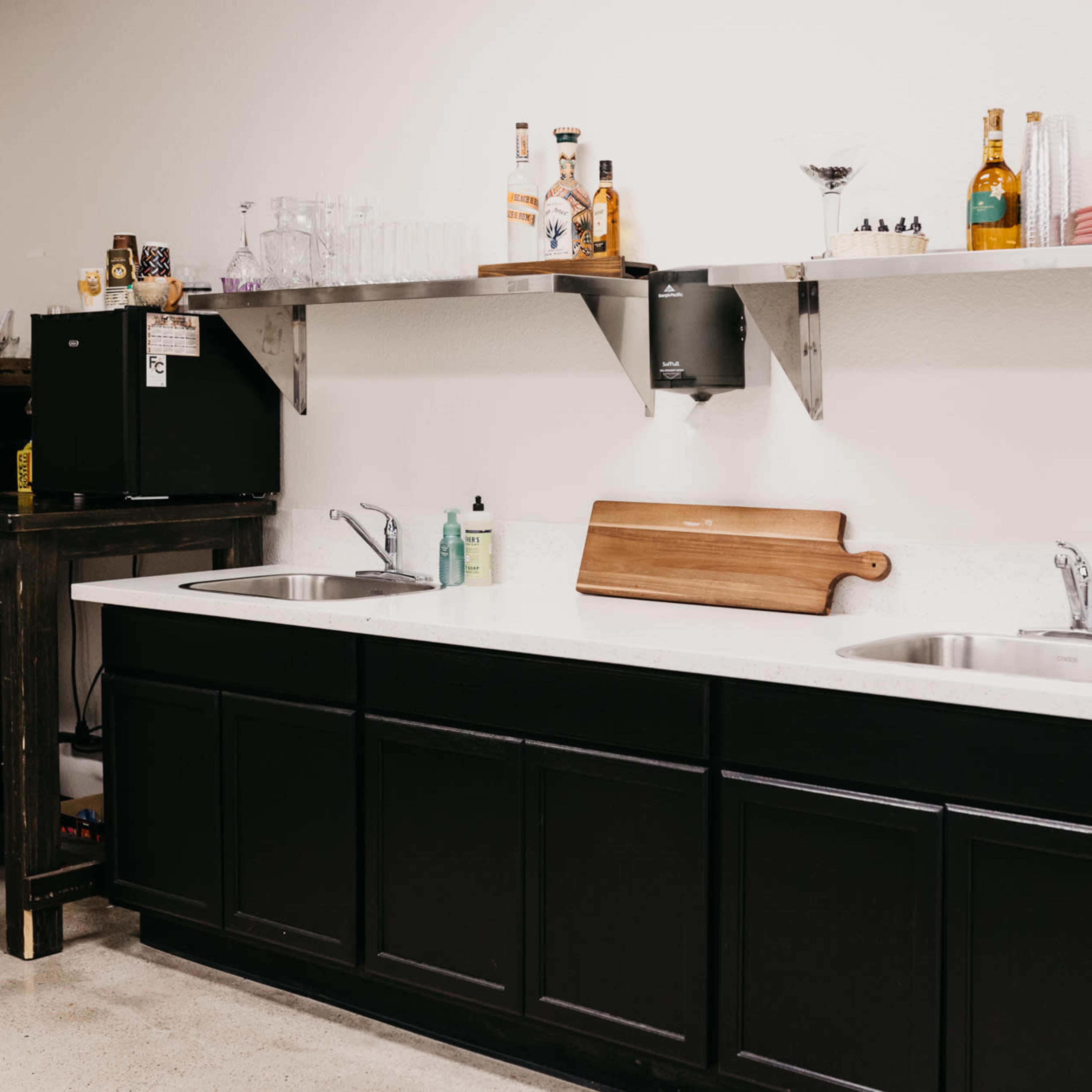 A kitchen area features a dark cabinet with a white countertop, two sinks, shelving with various bottles and glasses, and a microwave.