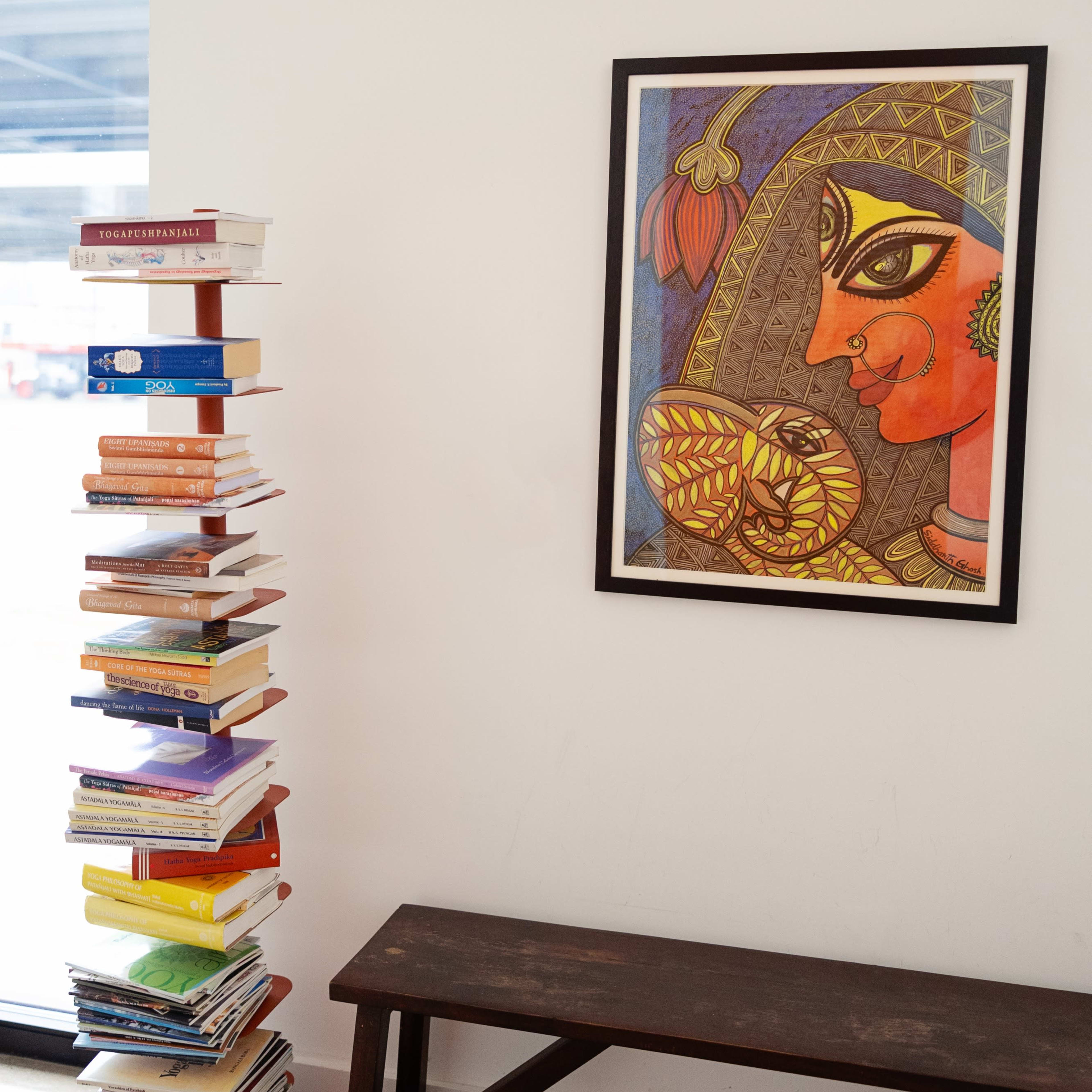 A tall stack of books stands next to a framed painting of a woman with decorative features on a wall.