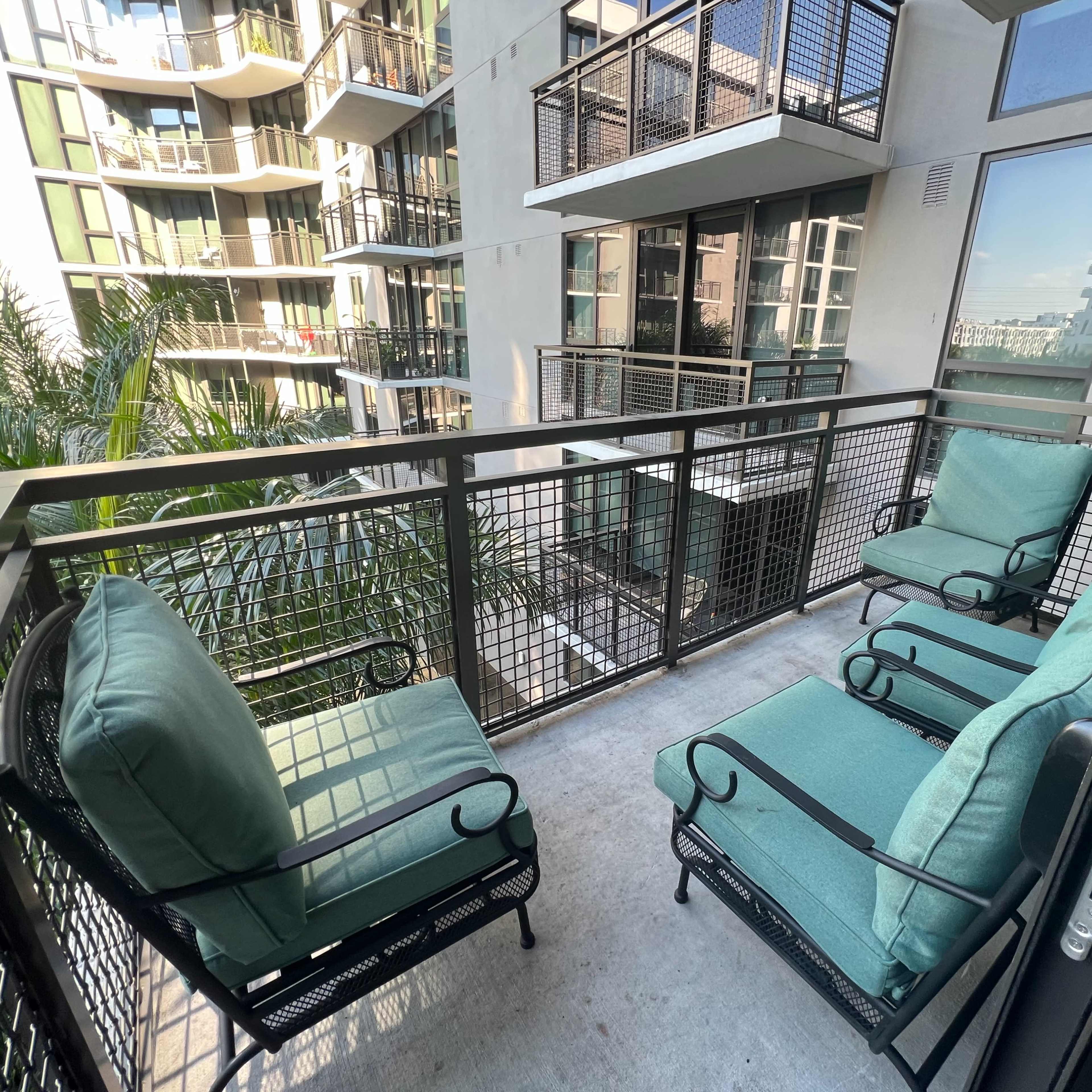 A balcony features four metal-framed chairs with green cushions and overlooks a modern apartment complex surrounded by palm trees.