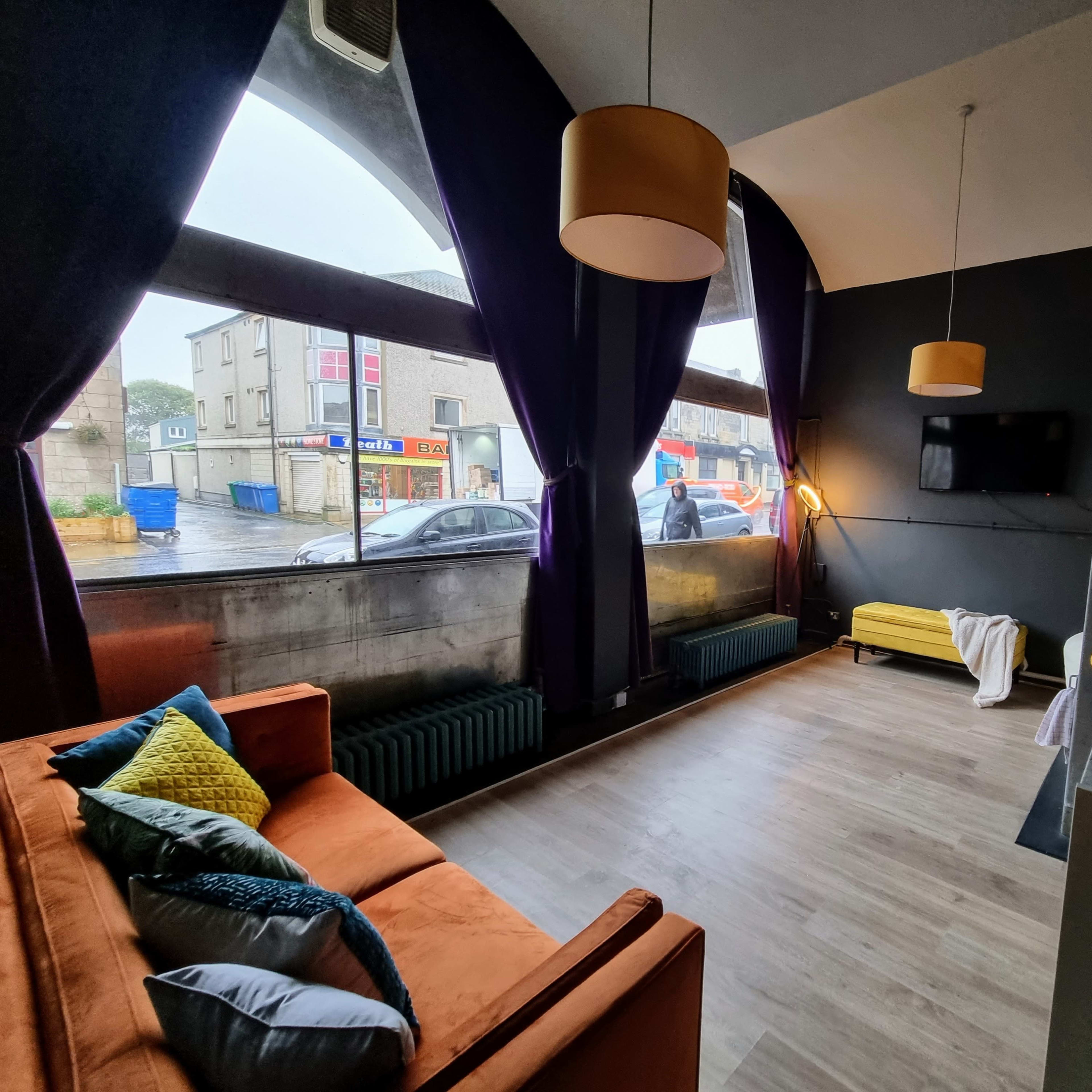 A modern interior of a room with large windows, a sofa, a kitchenette, and a view of a street outside.