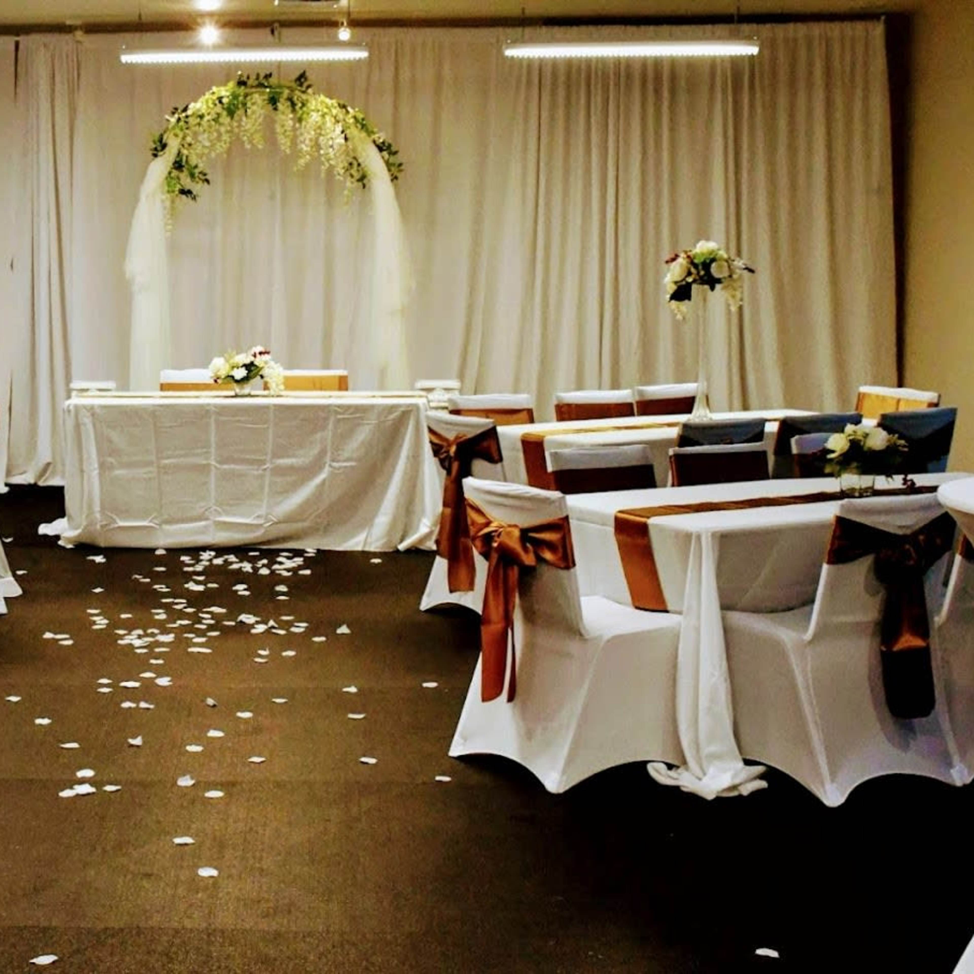 A wedding reception setup with covered tables, floral arrangements, and a decorated archway in a room with white drapery.