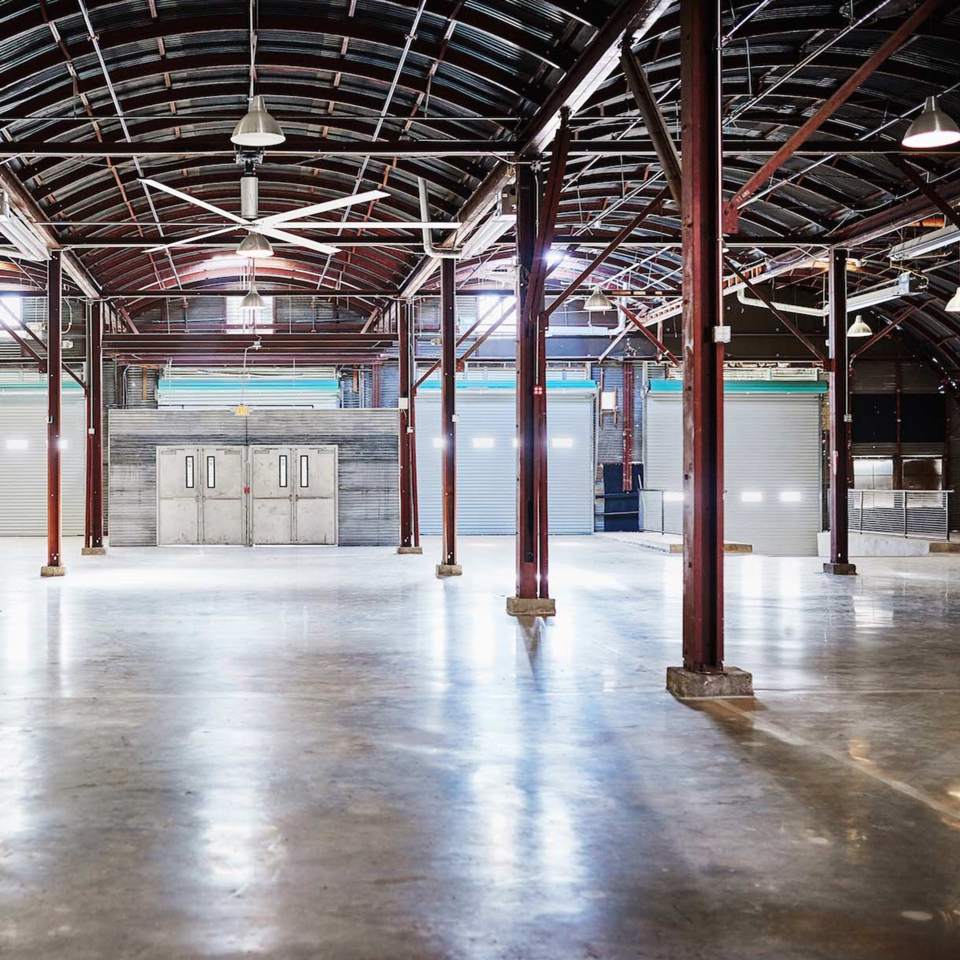 An empty industrial warehouse with large metal pillars, a polished concrete floor, and overhead lights illuminating the space.