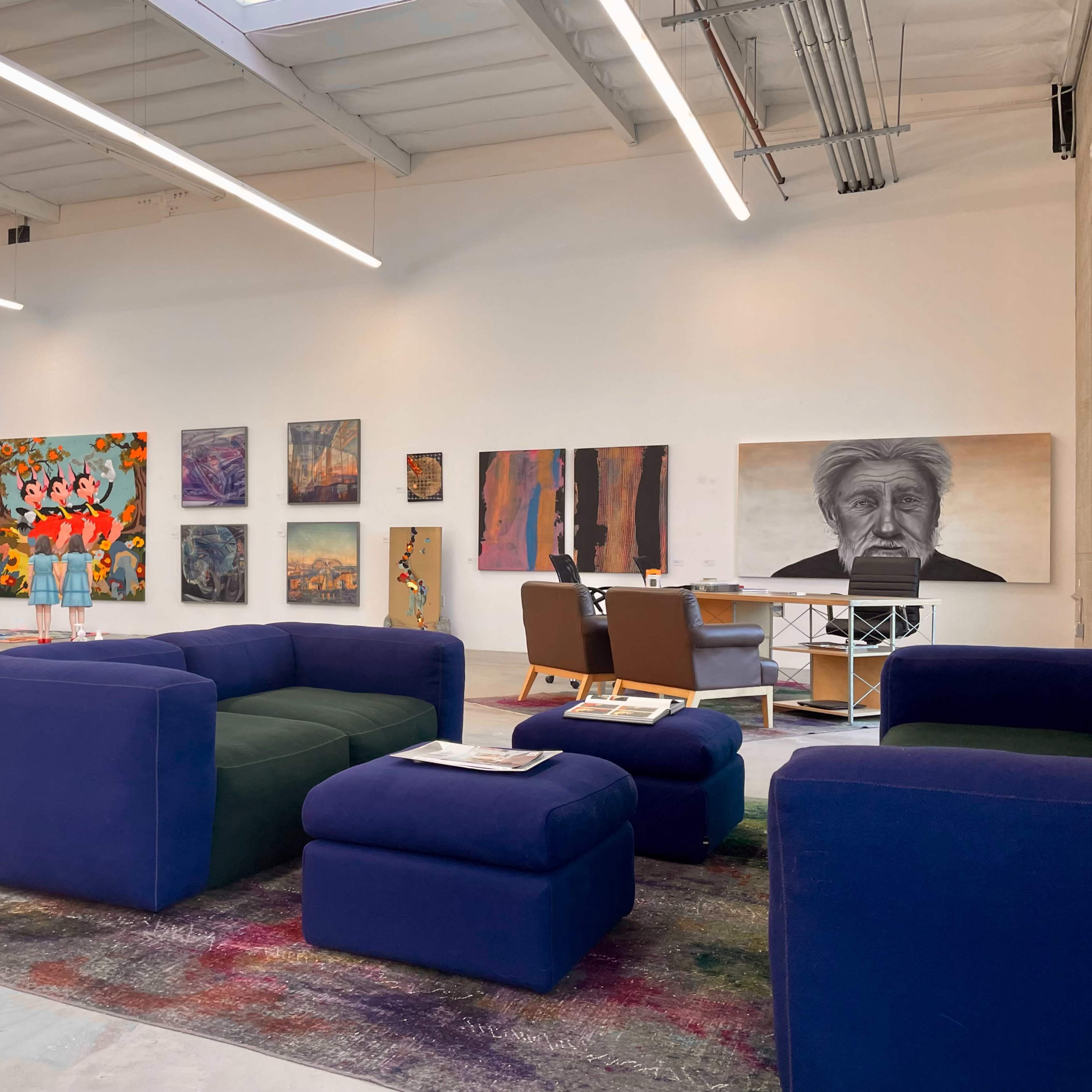 A spacious art gallery with colorful paintings on the walls and a seating area consisting of a blue couch and ottomans.