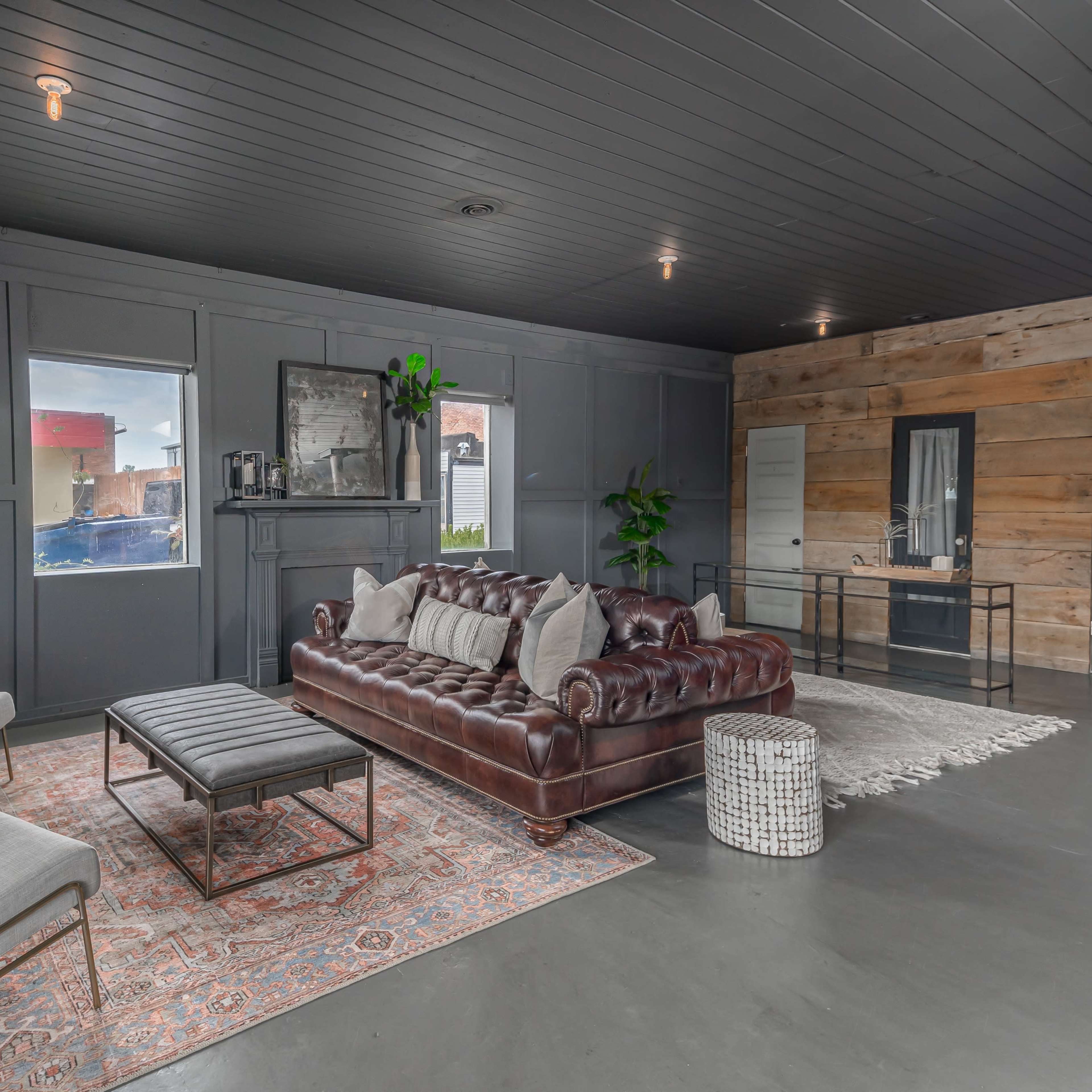 A modern living room with a dark ceiling, a brown leather couch, two light-colored chairs, and a rug on a concrete floor.