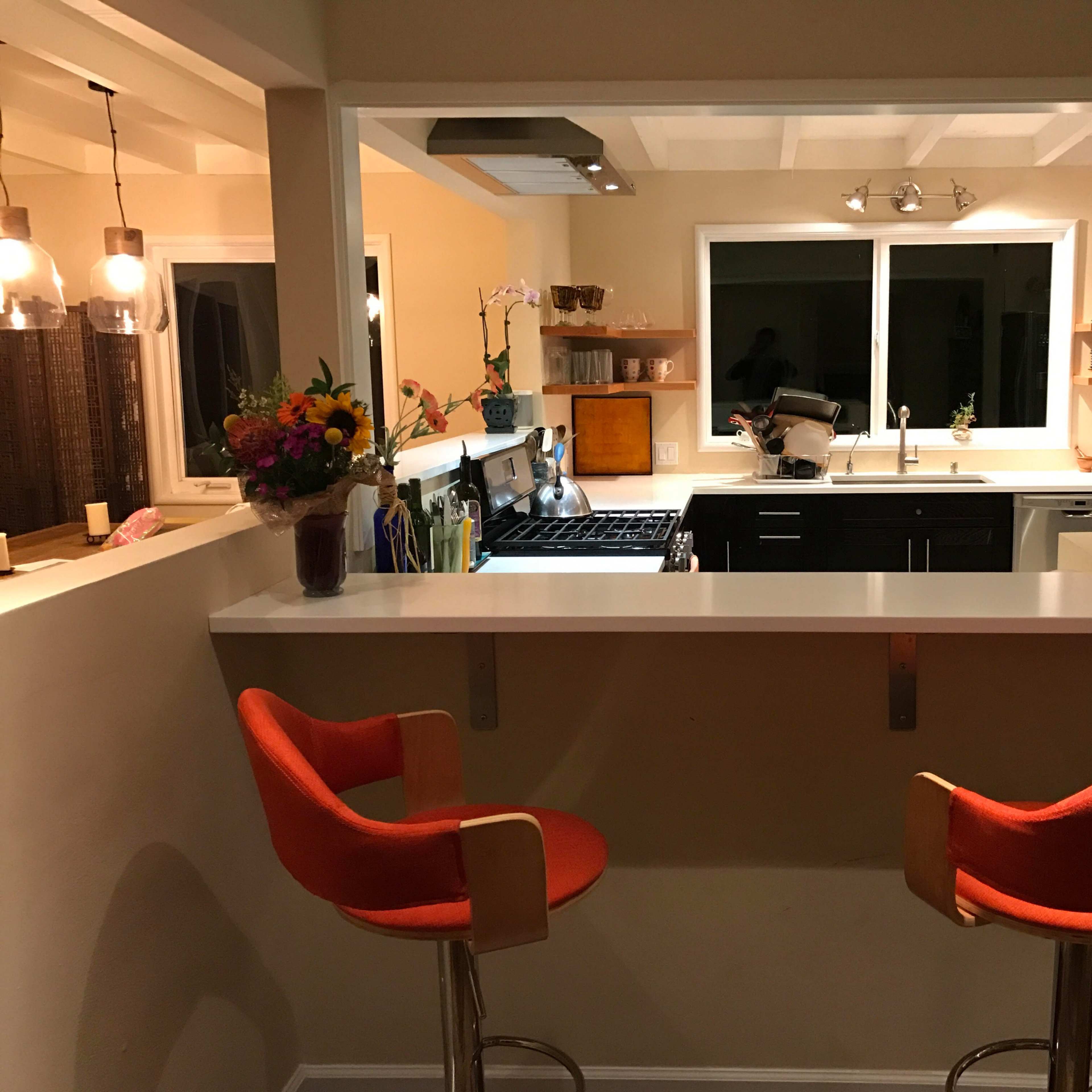 A modern kitchen features a bar with two orange stools, a gas stove, and a large window overlooking the sink area.