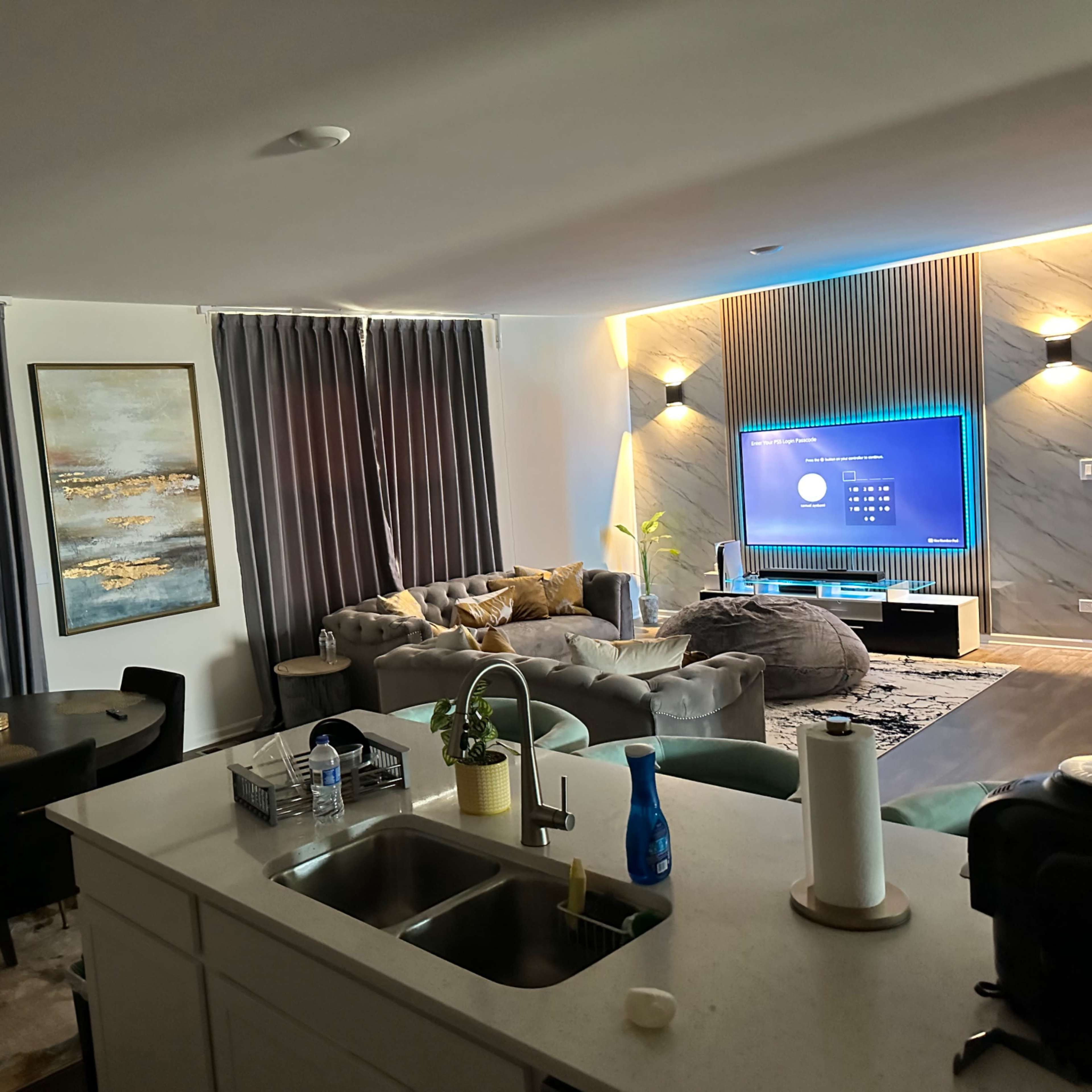 A modern living space with a kitchen in the foreground, a dining area with a black table, and a lounge area featuring a large couch and a wall-mounted TV.