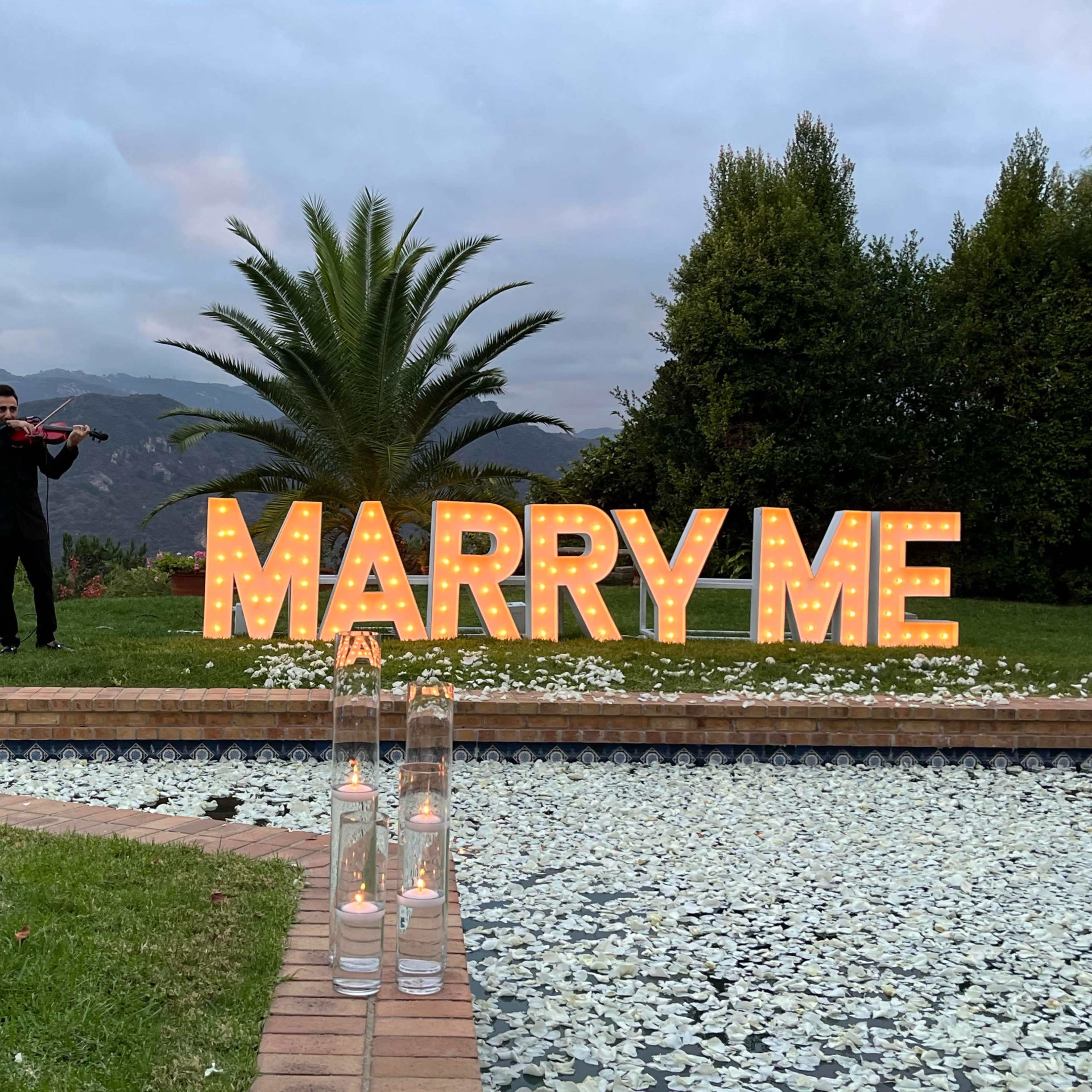 A man plays the violin in front of a large illuminated "MARRY ME" sign, surrounded by candles and a scenic backdrop of mountains and palm trees.