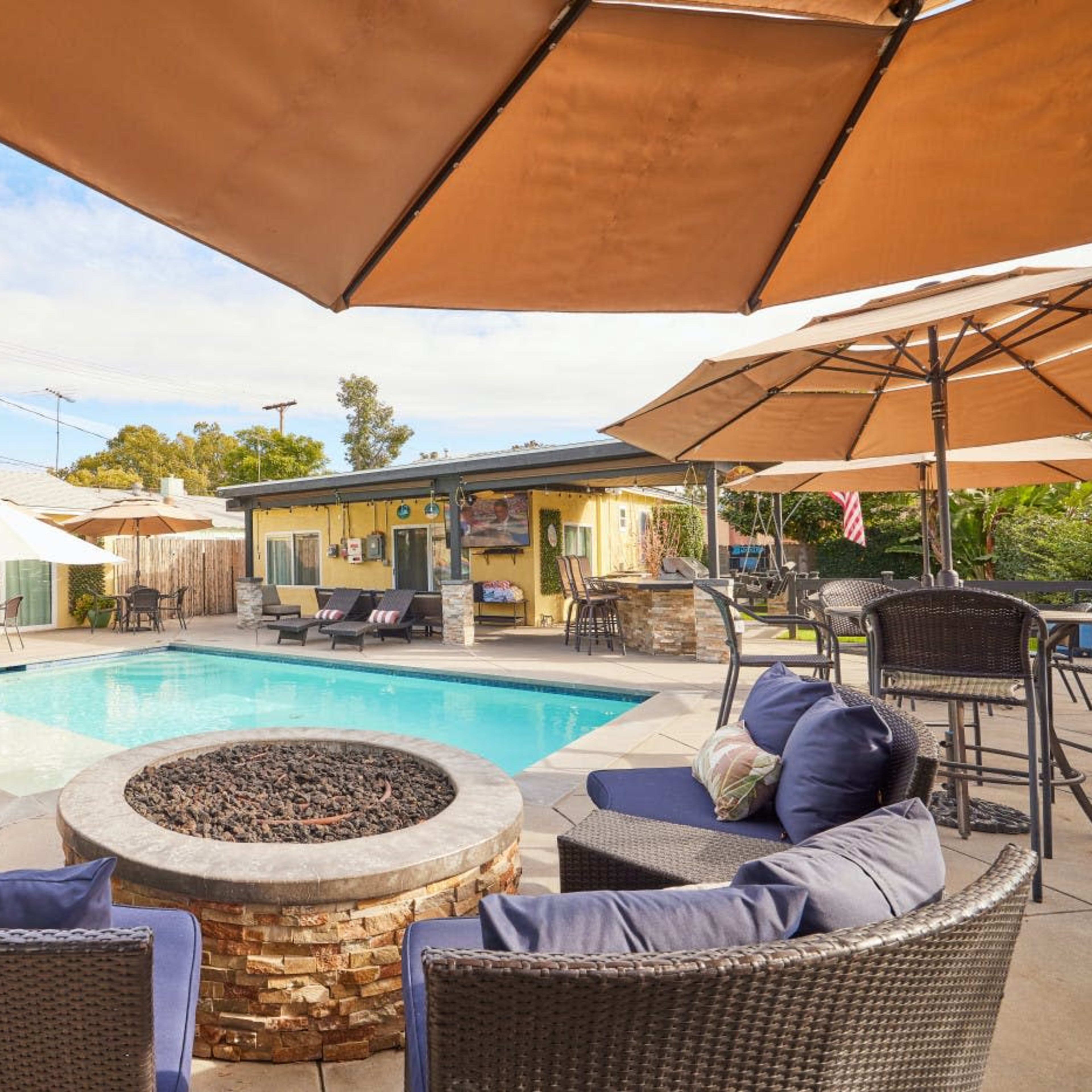 A backyard pool area with a fire pit surrounded by comfortable seating and several umbrellas for shade.