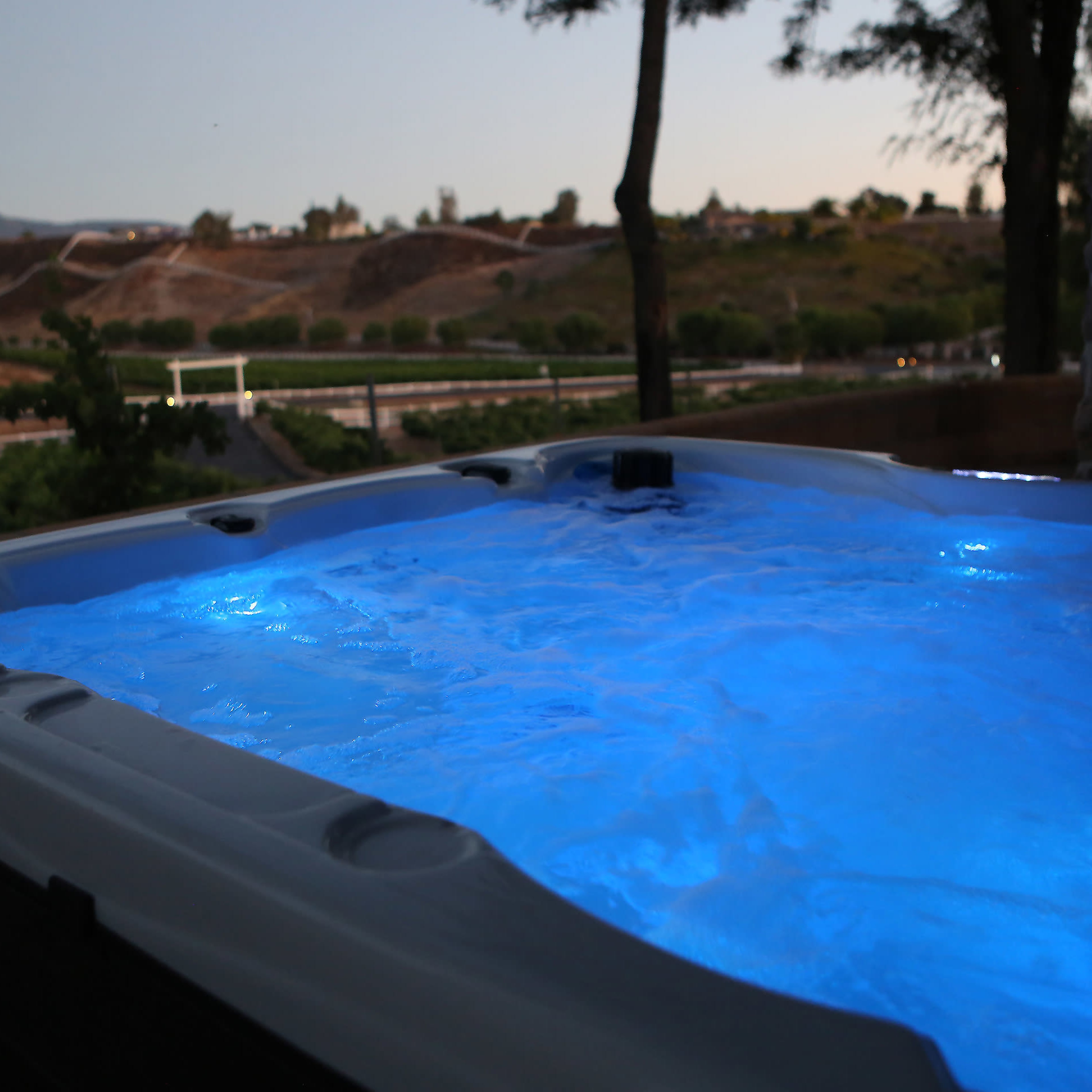 A hot tub with blue-lit water is situated outdoors, surrounded by trees and overlooking a hilly landscape at dusk.