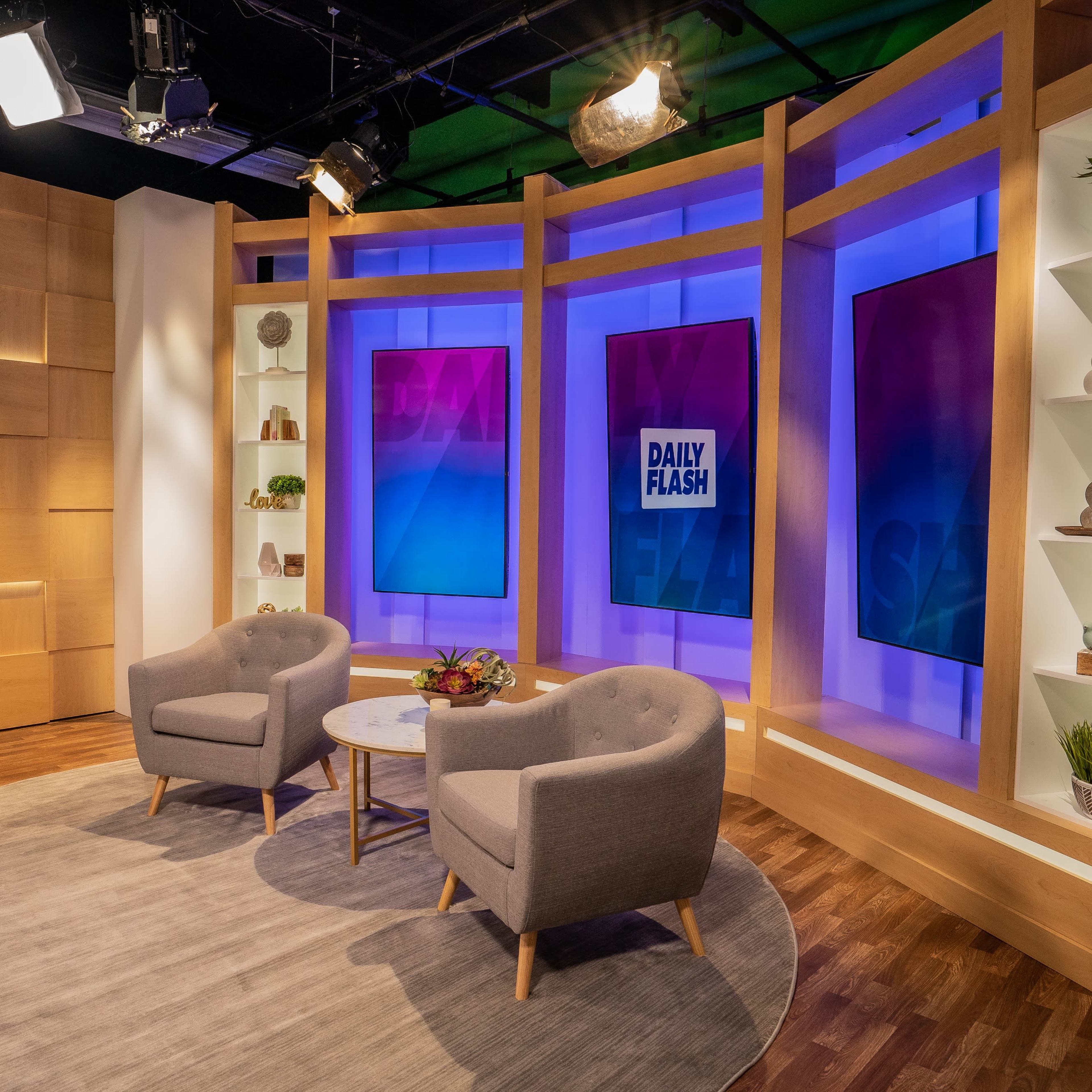 A television news studio set with two gray chairs and a round table, illuminated by colorful backdrops and a screen displaying "DAILY FLASH."