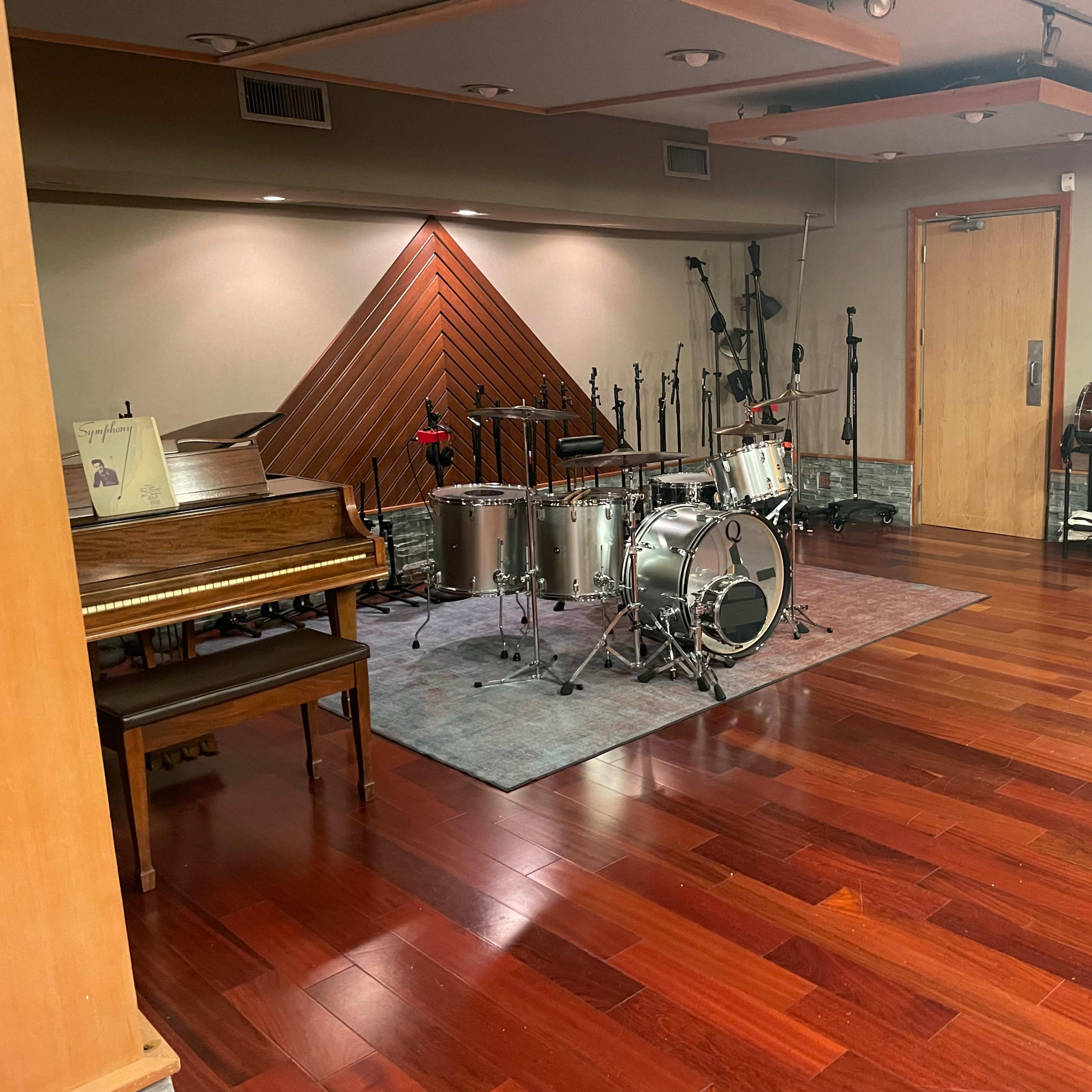 A music studio with a grand piano on one side and a drum set on a rug in the center, surrounded by microphones and various instruments on the walls.