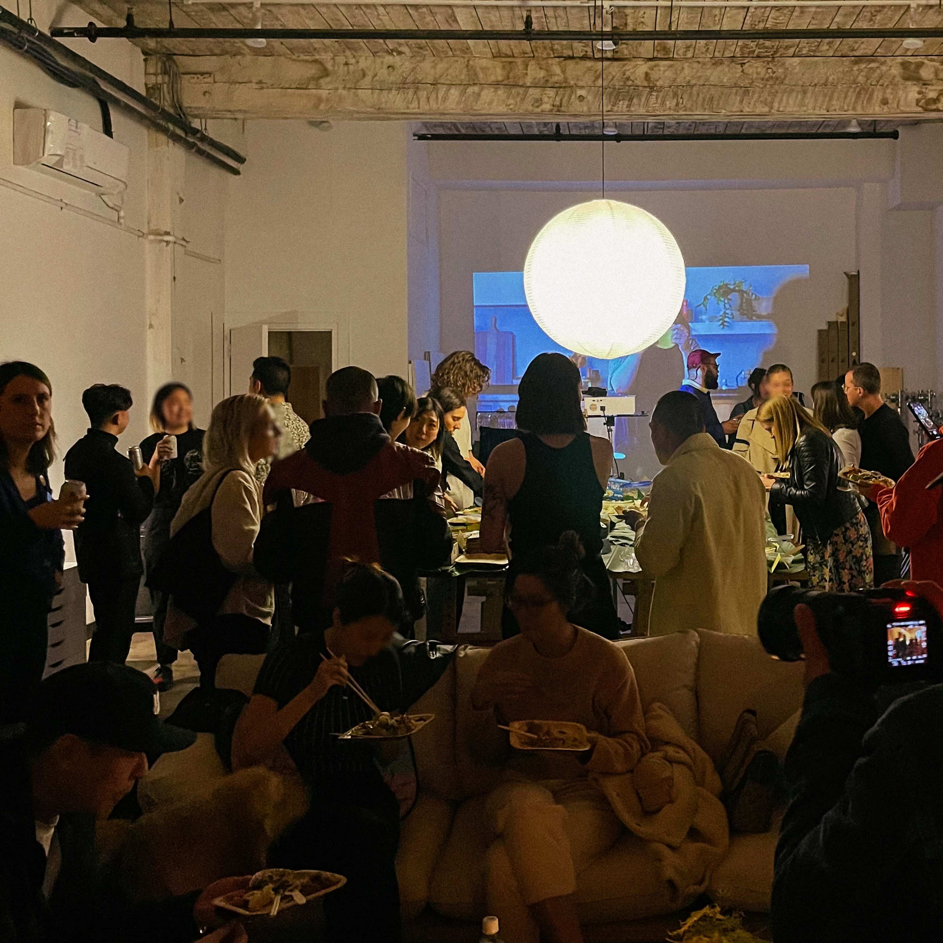 A crowded indoor event features guests socializing and dining in a well-lit space, with a large round light fixture hanging overhead.