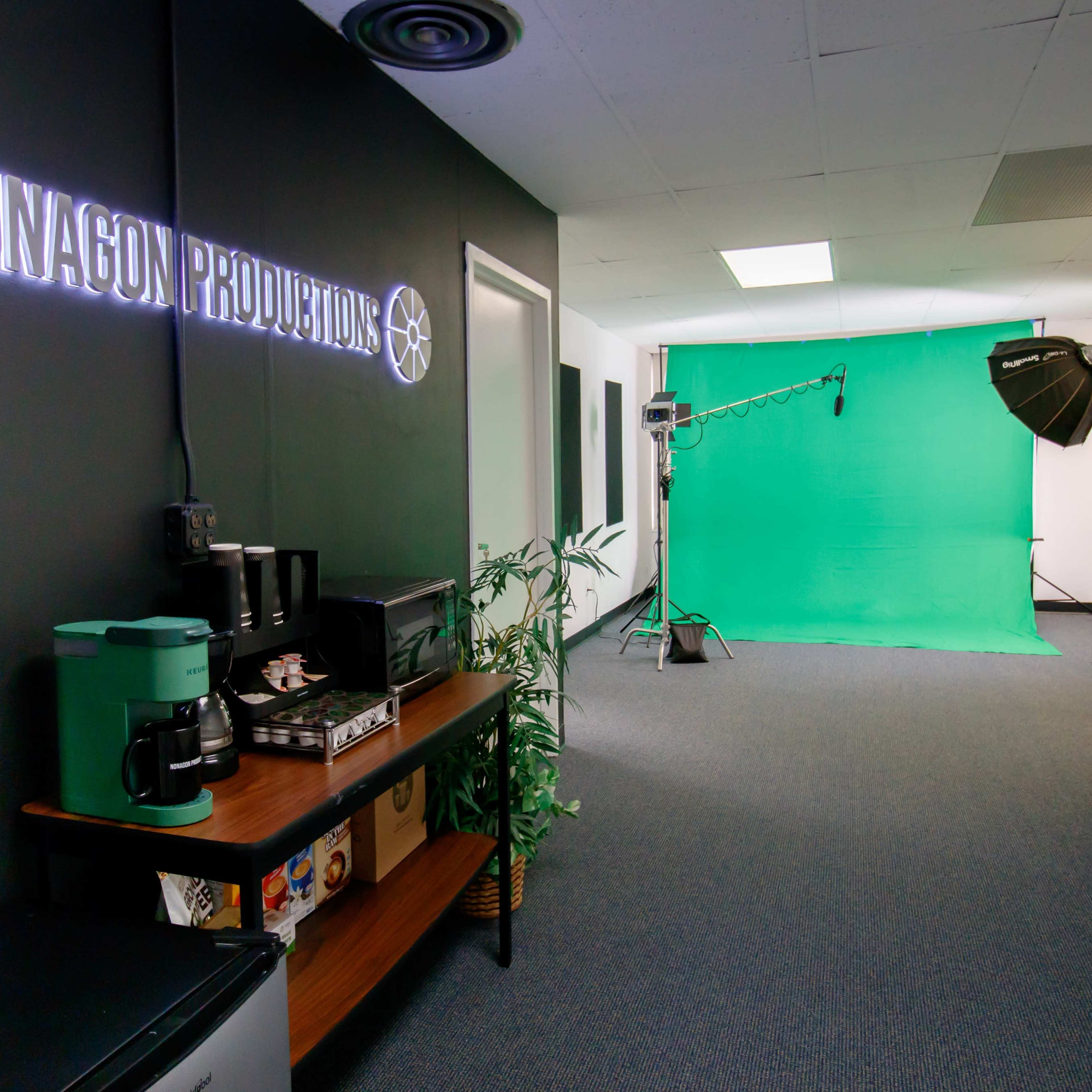 An office space with a green screen setup, a coffee station on a wooden table, and lighting equipment arranged for production purposes.